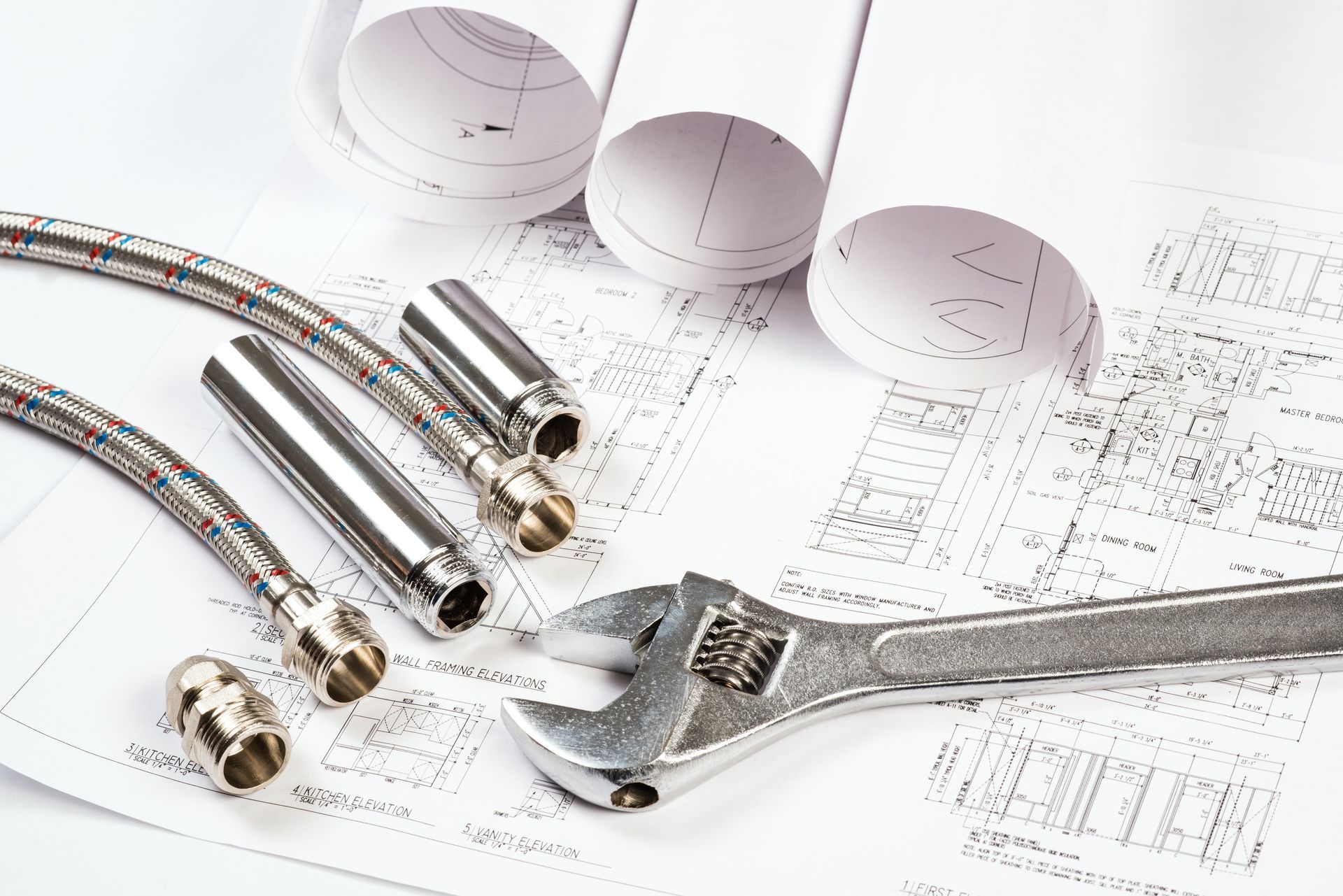 Outils et plans de plomberie : tuyaux flexibles, raccords métalliques, clé à molette et plans architecturaux roulés sur une surface blanche.