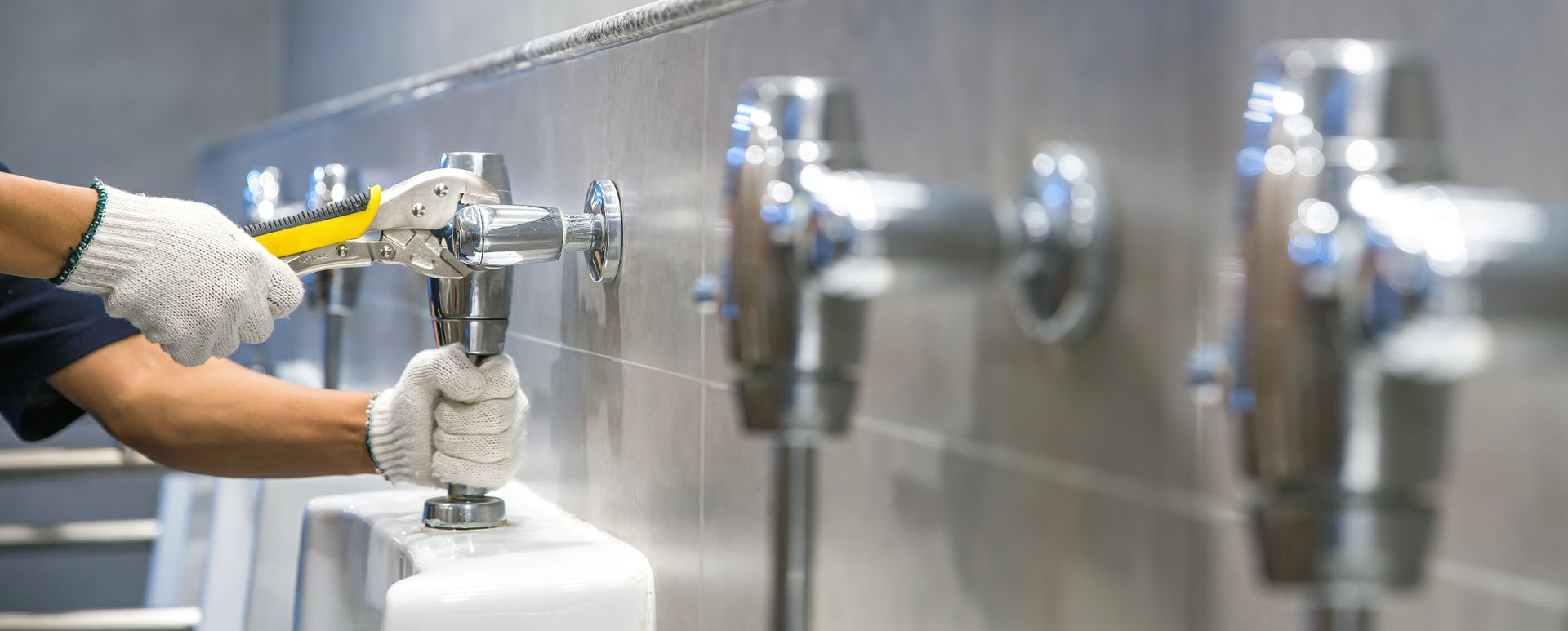 Un plombier, portant des gants, répare un urinoir dans des toilettes publiques, à l'aide d'une clé.