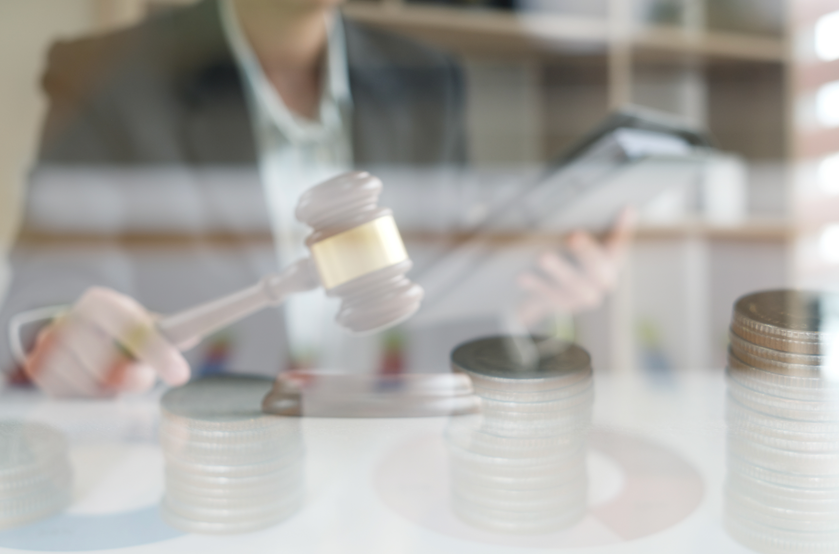 A person holding a gavel over stacks of coins and documents; blurred background.