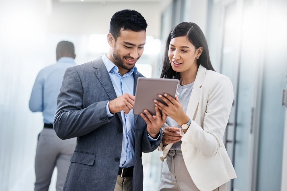 Two professionals look at a tablet in a hallway; man points at screen, woman smiles, another person walks behind them.