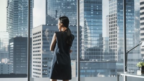 Woman in a dark dress with arms crossed, looking out a large window at a cityscape.