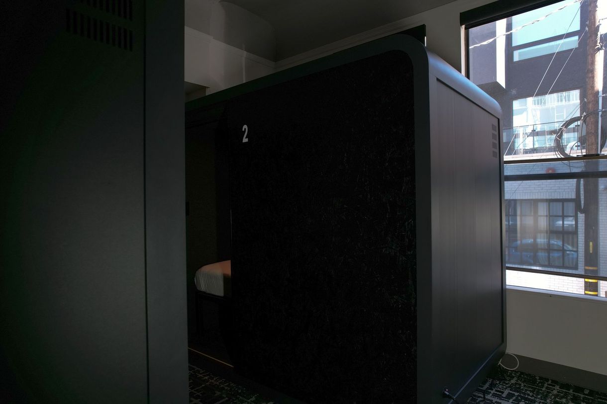 A soundproof booth with a dark, textured front stands in a room with a window showing a cityscape.