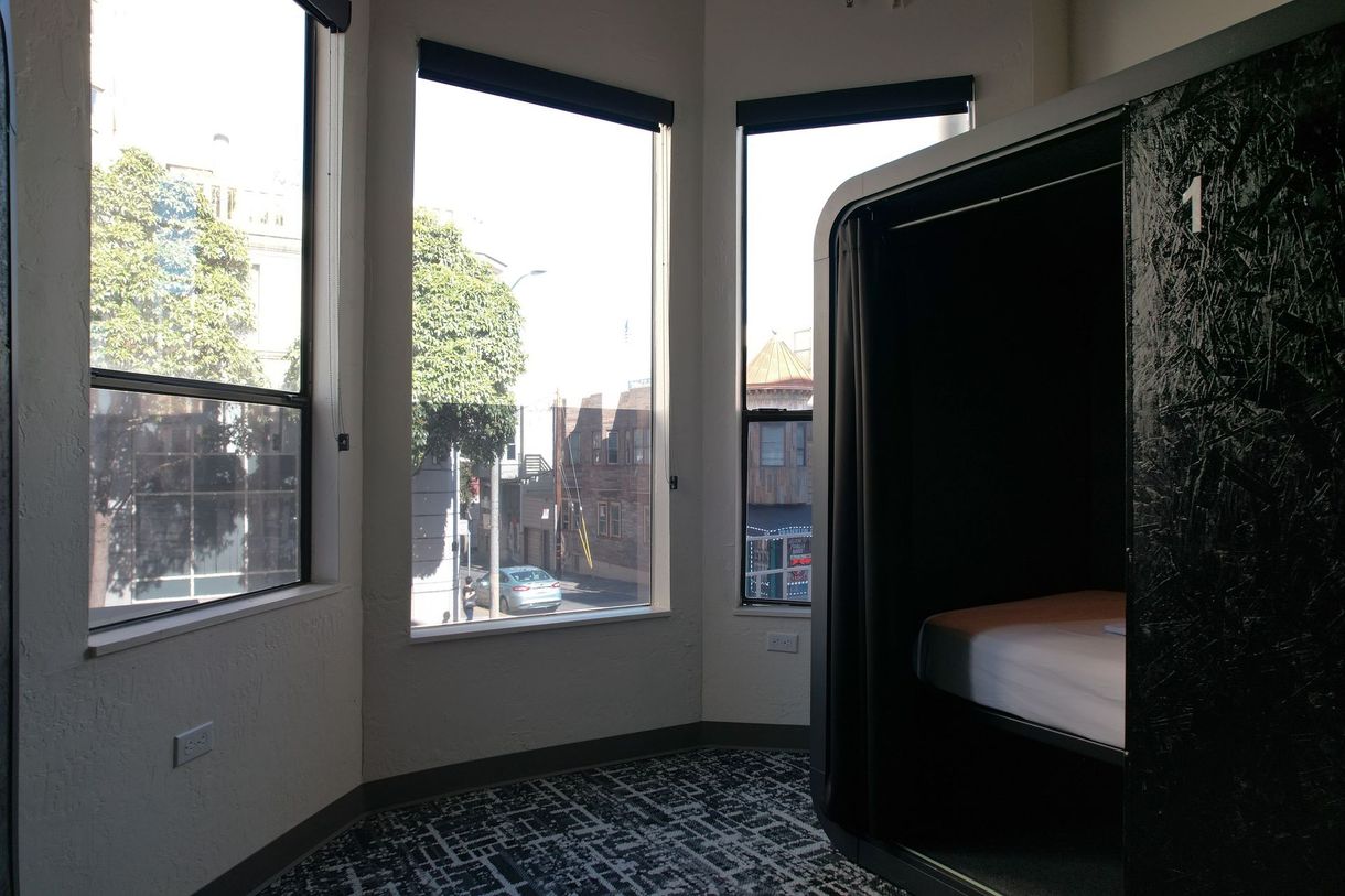 A capsule hotel room with a bed inside, next to large windows overlooking a street.