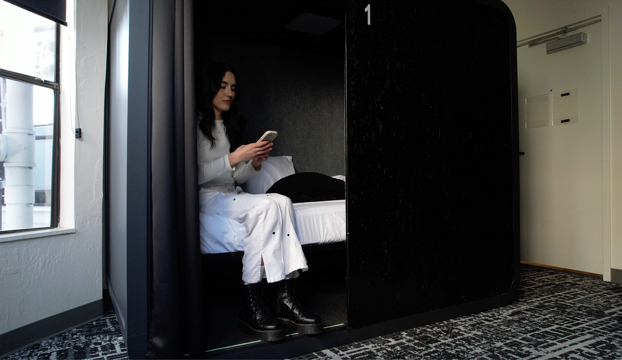 Woman in a black pod using a phone, white pants and a white shirt. The pod has a bed.