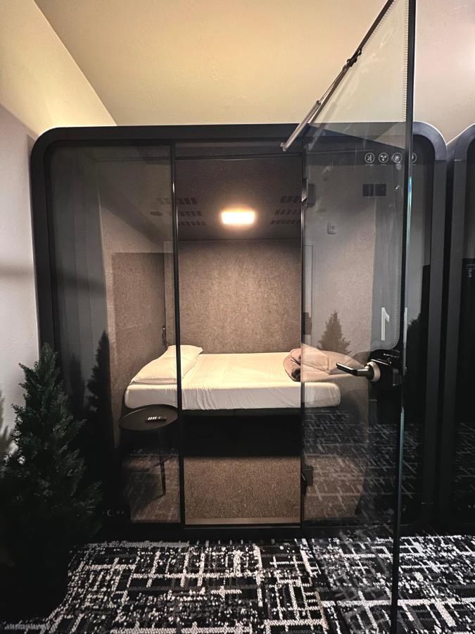 Bedroom pod with glass doors, bed, and overhead light. Dark gray walls, floor, and trim.