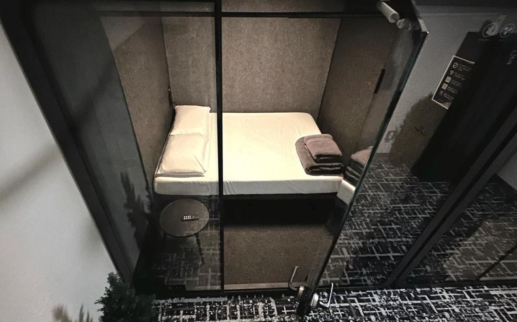 Small, enclosed modern bedroom with a bed, small side table, and towels. Black and grey tones dominate.