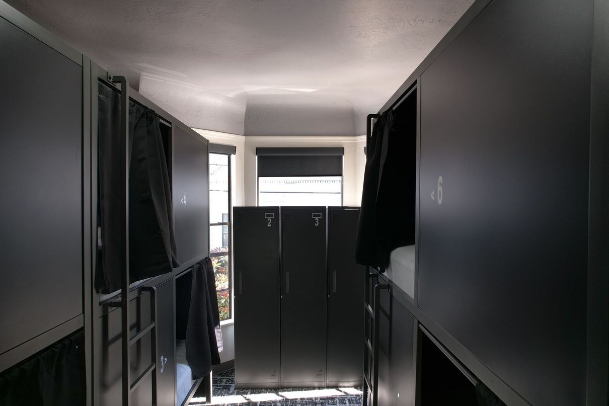 Narrow hostel room with bunk beds and lockers; a window is visible at the end of the hall.
