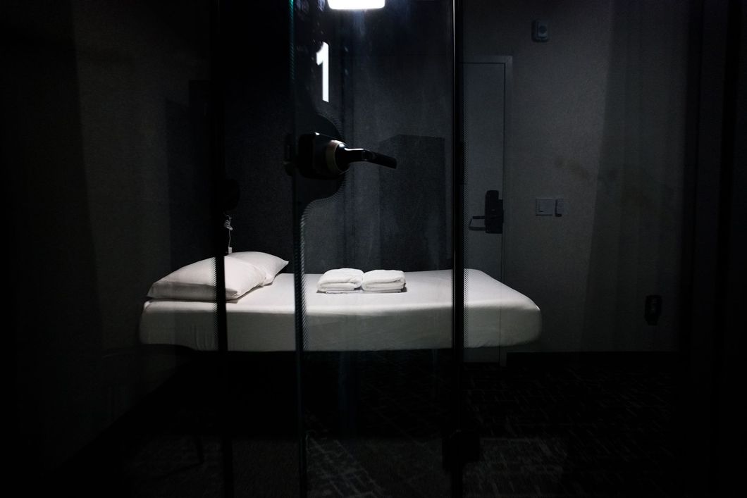 Dark room with single bed inside a glass enclosure, lit by overhead light.