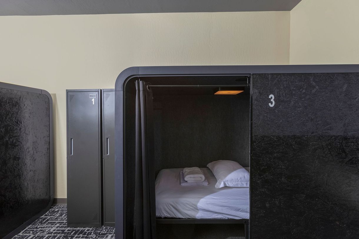 Capsule hotel room, black with bed, privacy curtain, small light, gray lockers.