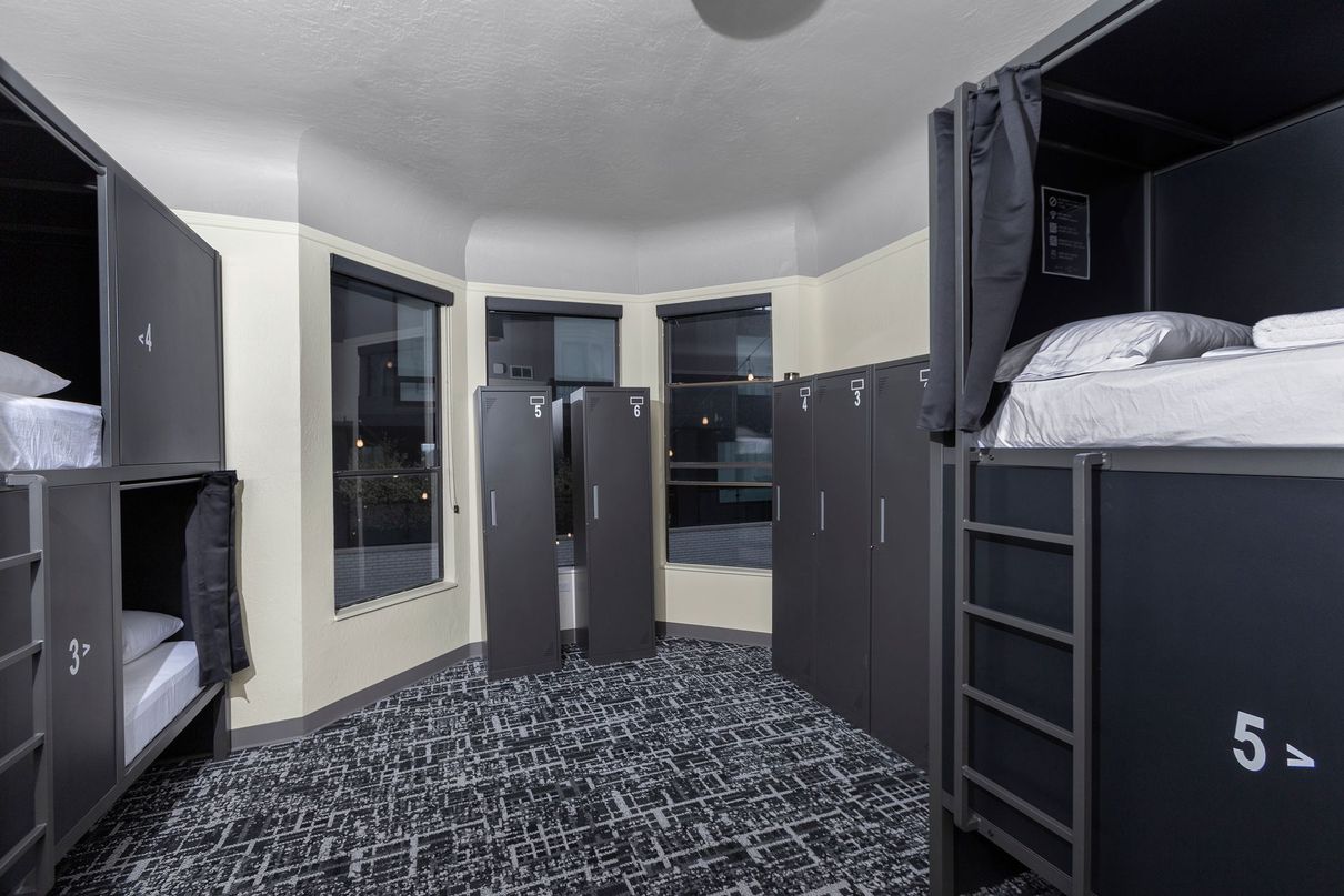 A hostel room with bunk beds, lockers, windows, and patterned carpet.