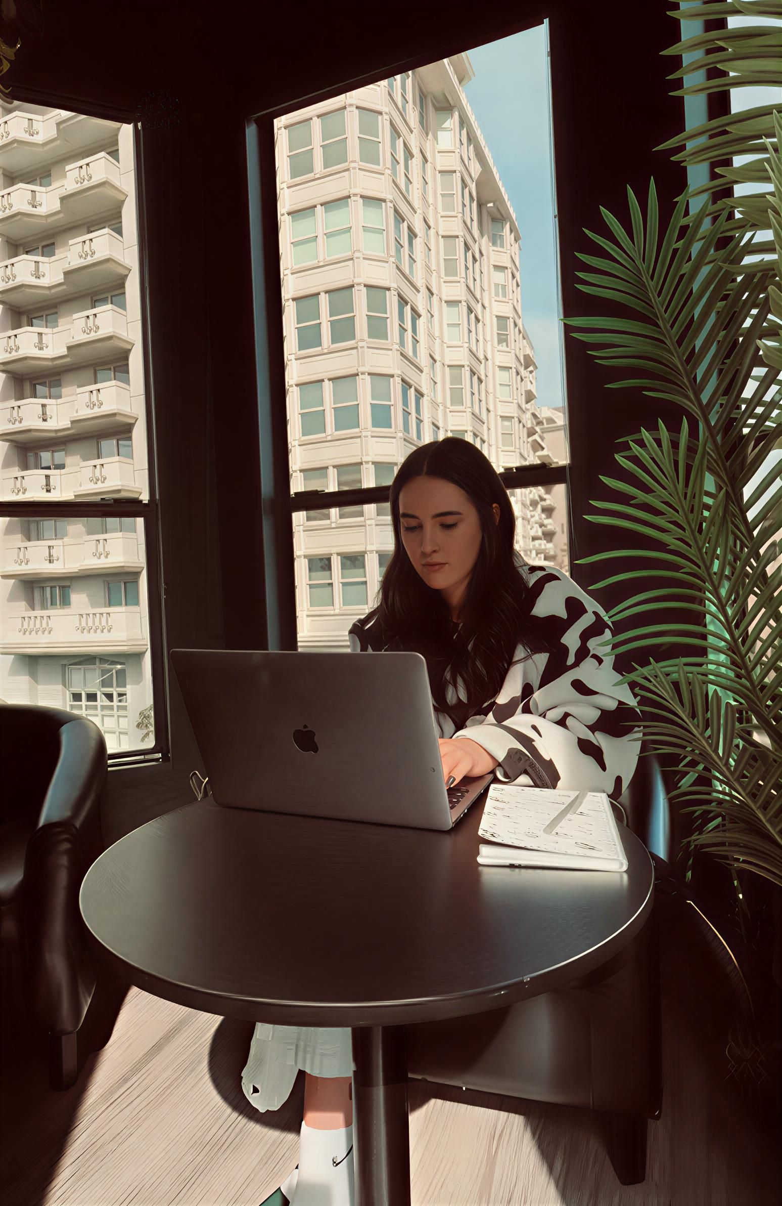 Woman working on laptop at a table near a window overlooking a city.