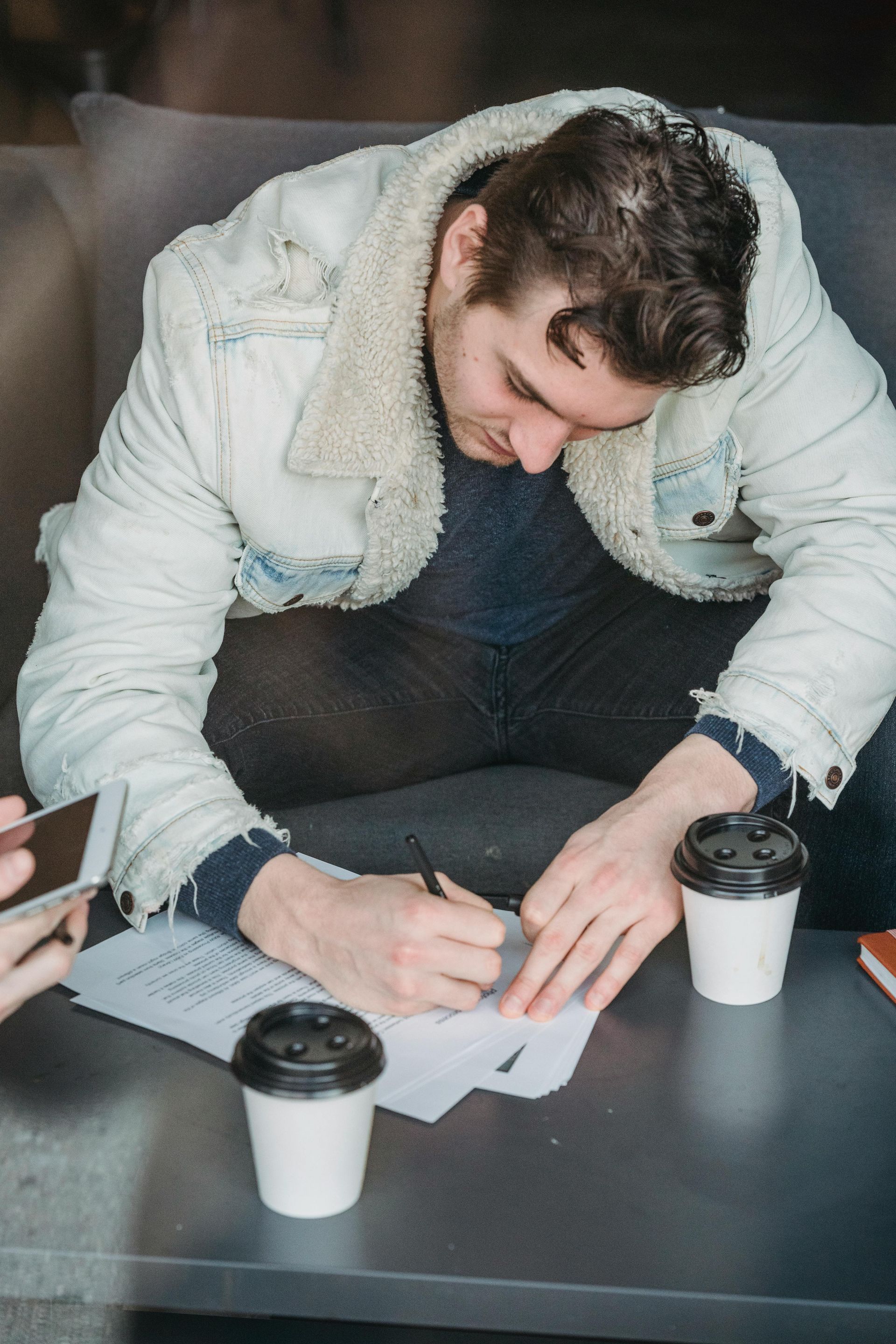 Man writing on paper at a table, wearing a white denim jacket, with two coffee cups.