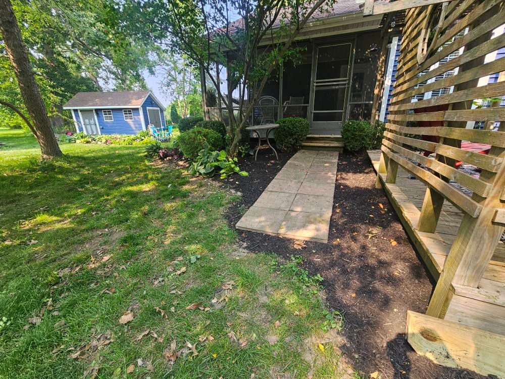 Wooden walkway through a shaded backyard garden beside a house, with a blue shed in the distance