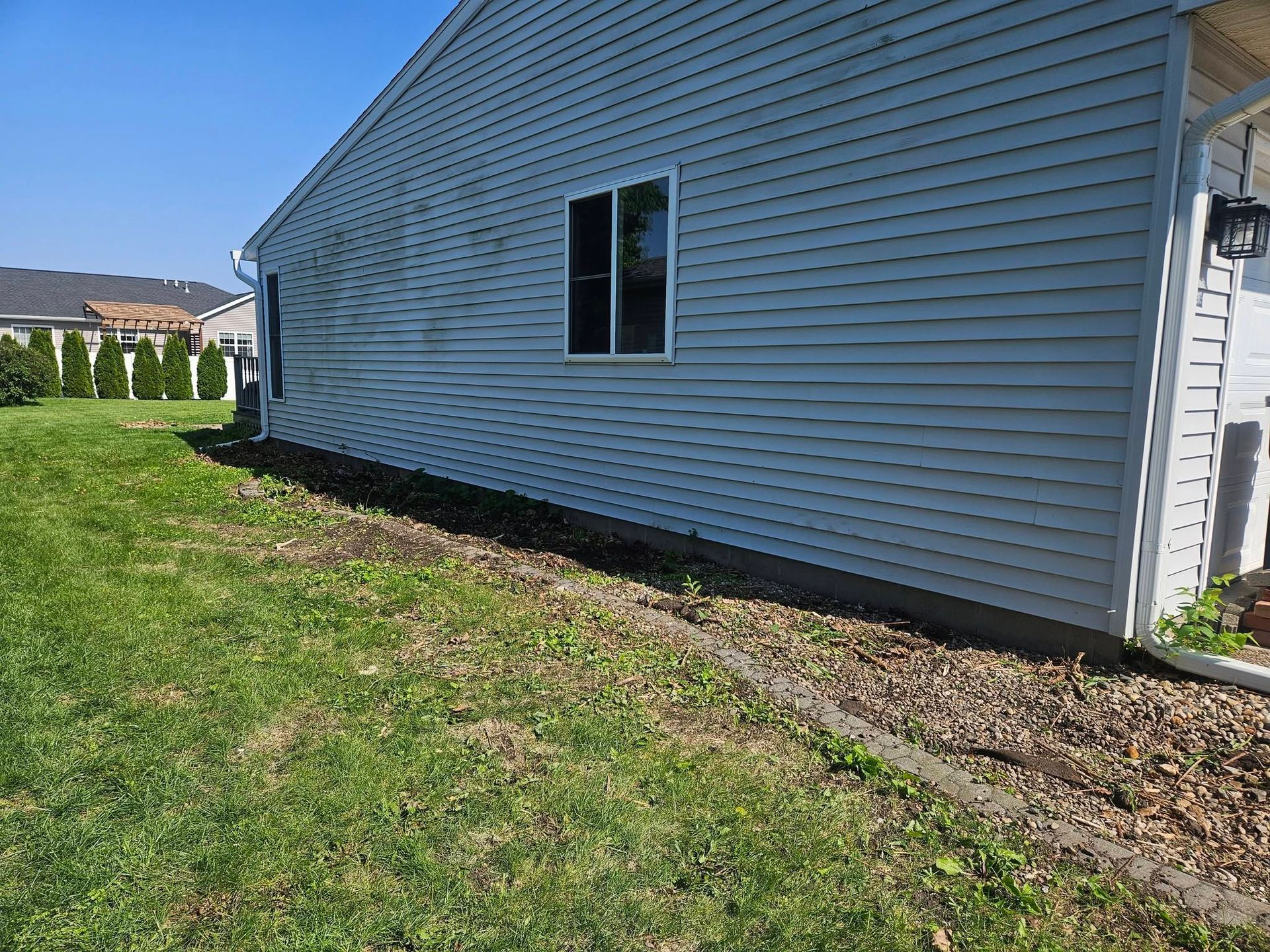Side view of a white vinyl-sided house with a window, next to a grassy yard and dirt border.