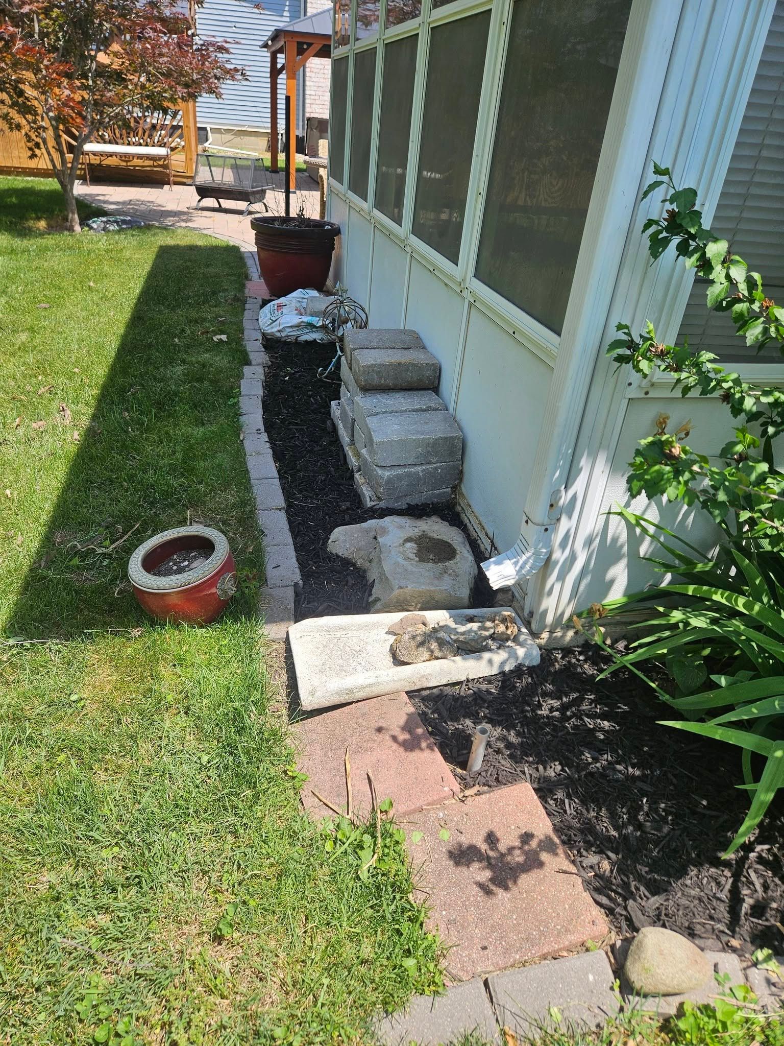 Backyard side yard with window wells, stacked pavers, and a narrow gravel strip beside a house