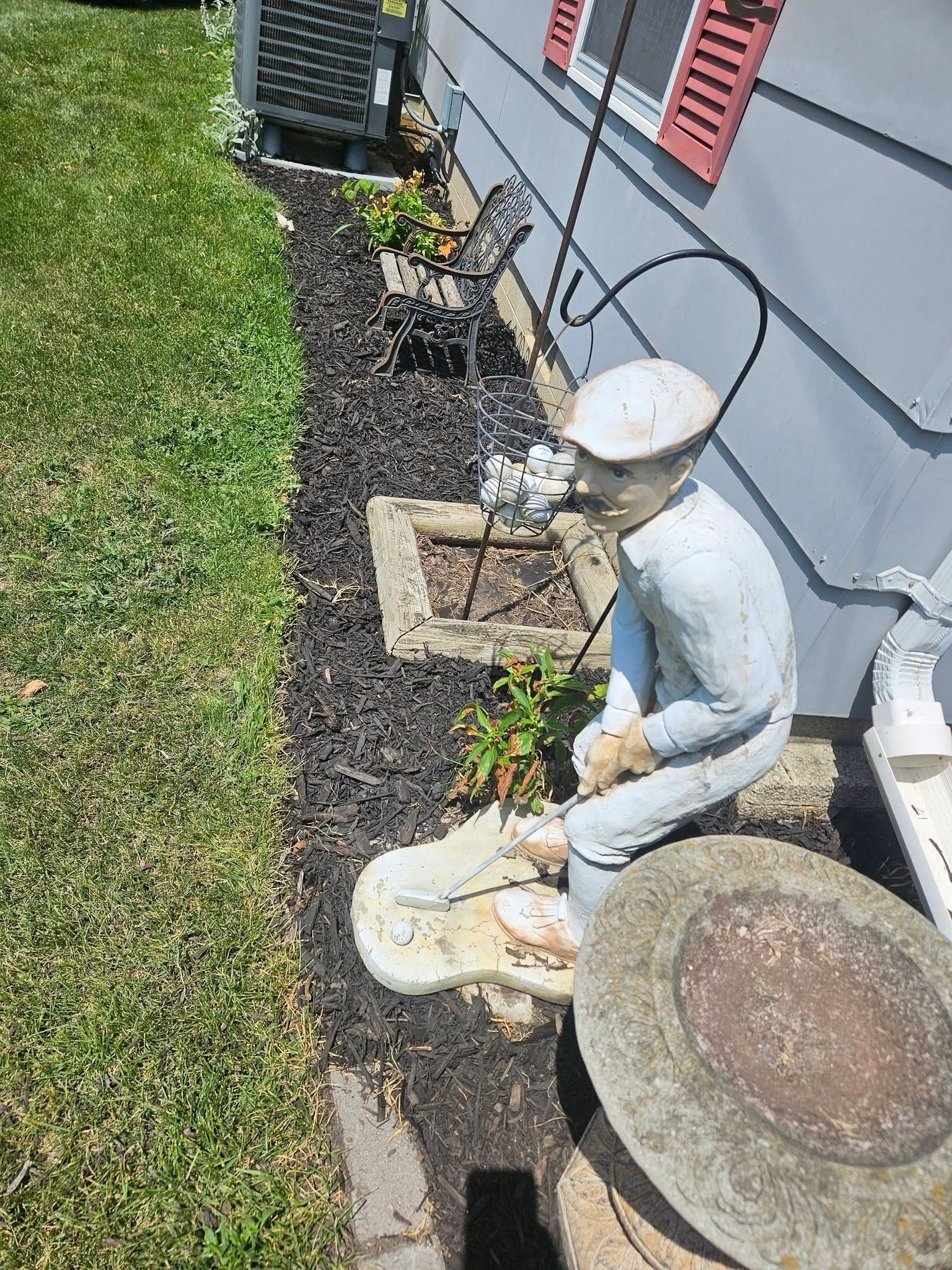 Small white garden figurines beside a house wall and vent, next to a large round stepping stone and mulch bed