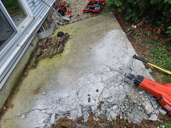 Cracked concrete patio beside a house, with a red power tool and debris on the ground.