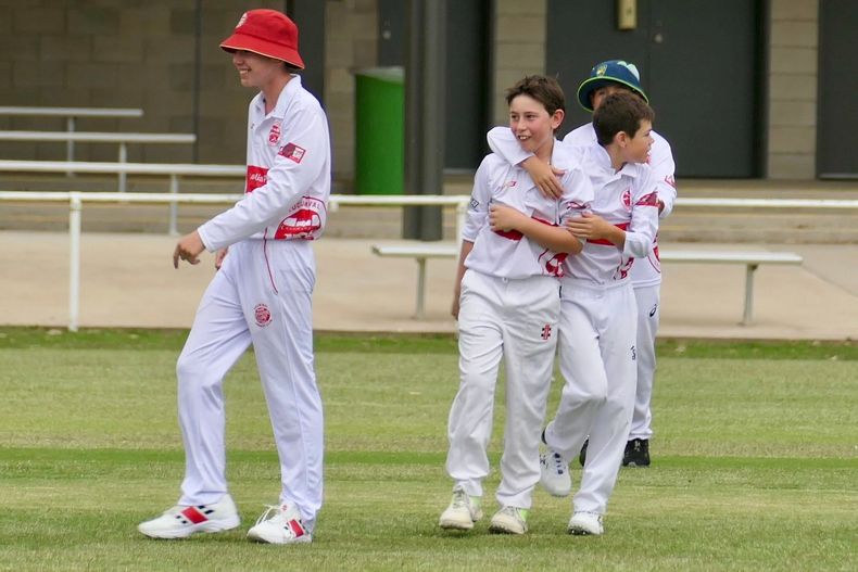 Cricket players in white uniforms standing on a grassy field, one wearing a red hat