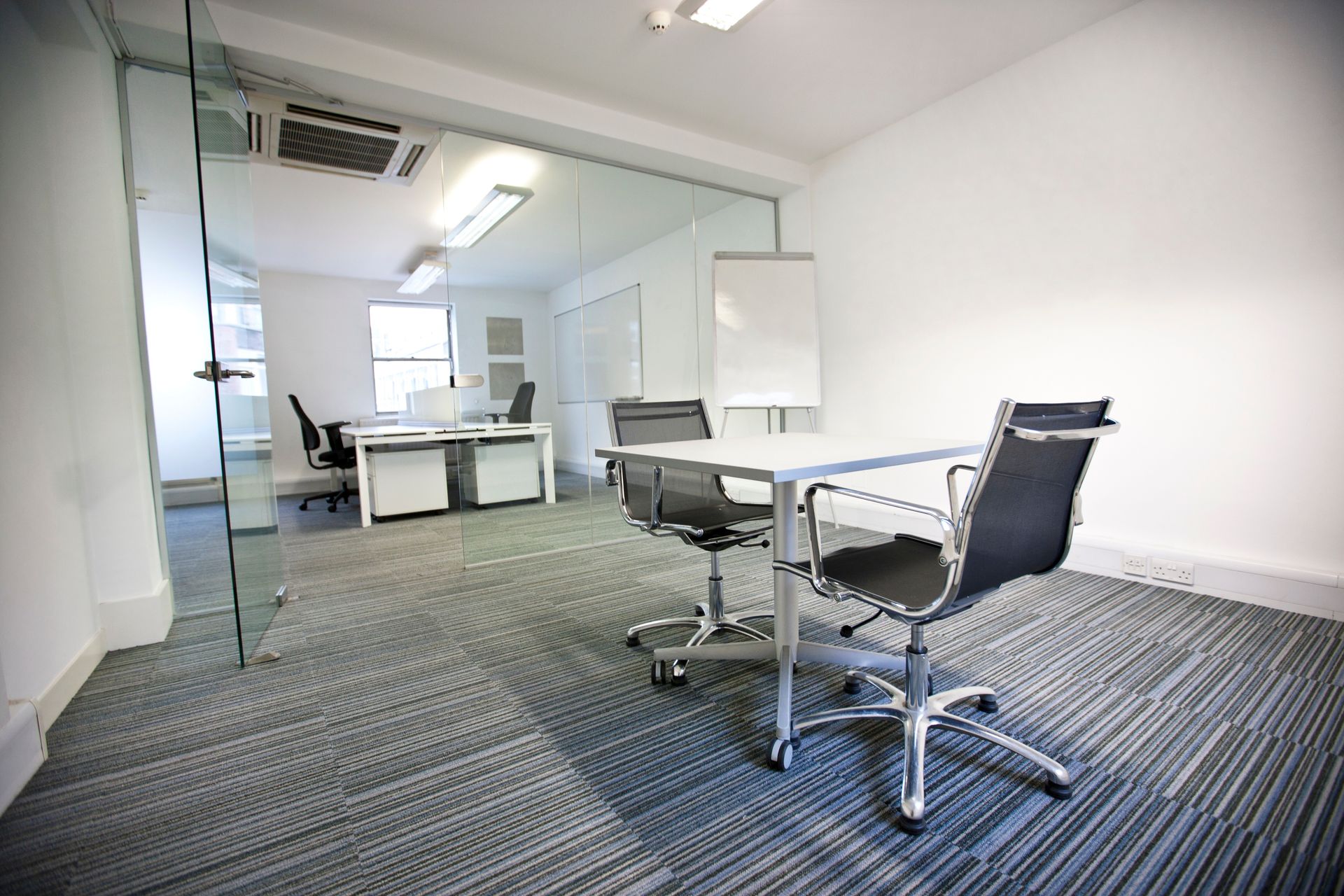 Empty modern office with desks, chairs, and carpeted floor, viewed through a glass wall