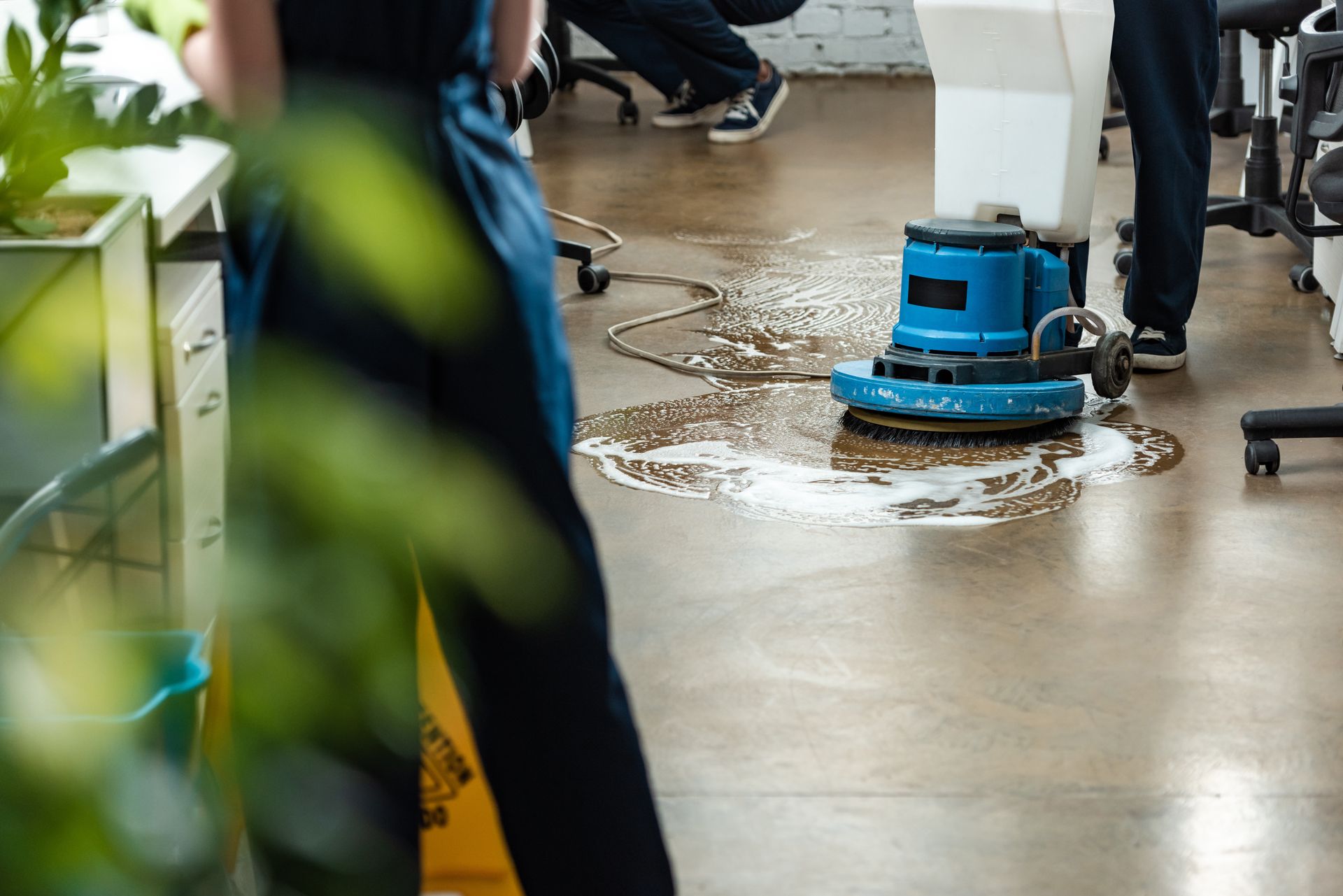 A blue floor scrubber cleans a wet floor in a busy indoor workspace.