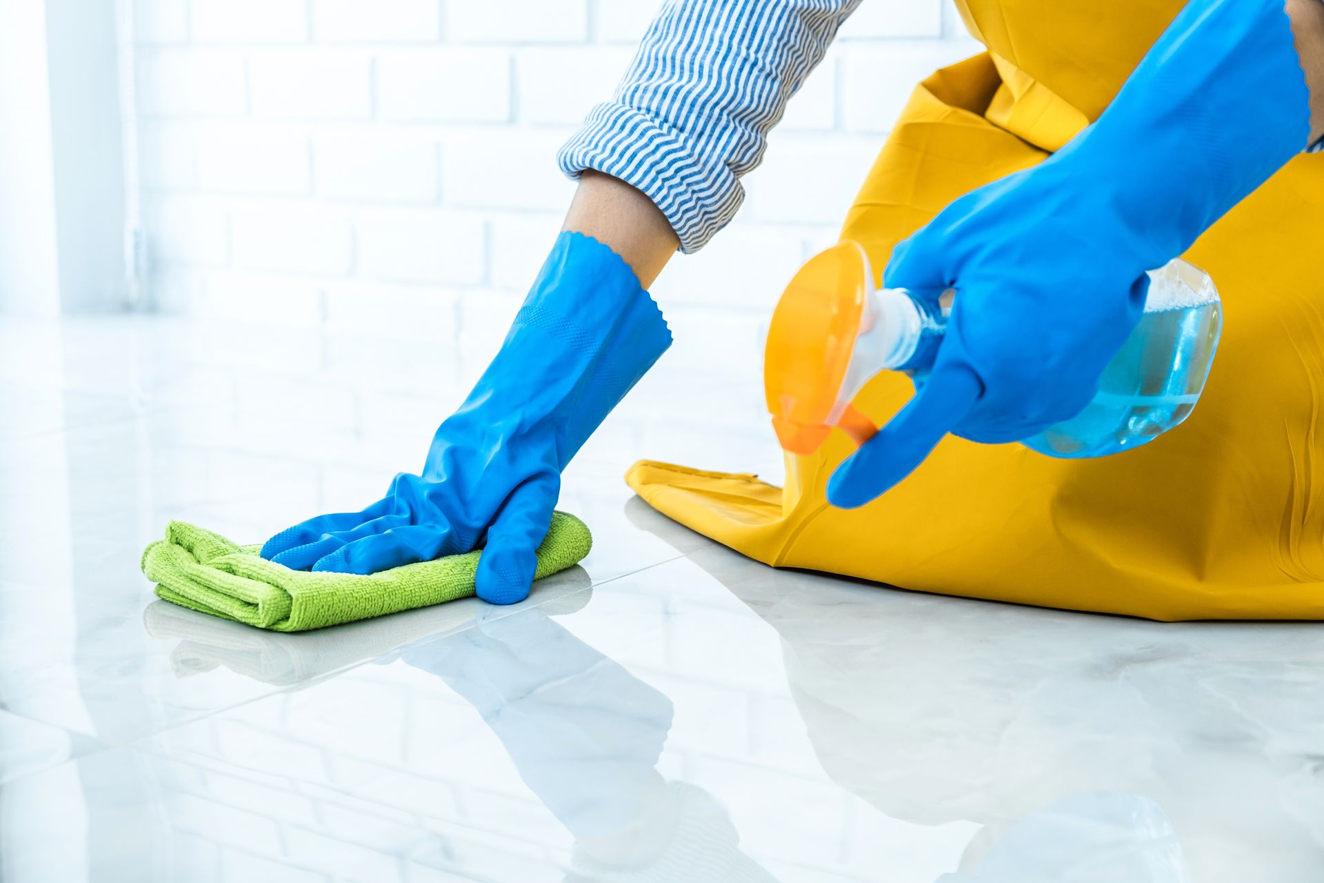 Person cleaning a tiled floor with a green cloth and spray bottle, wearing blue gloves and a yellow apron