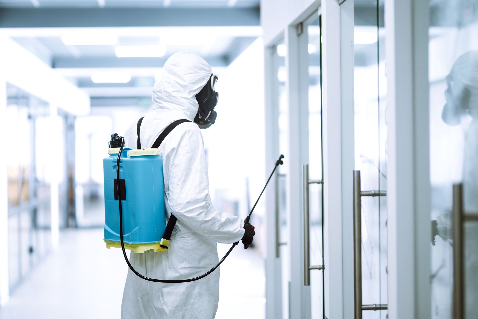 Worker in white hazmat suit spraying disinfectant in a bright hallway