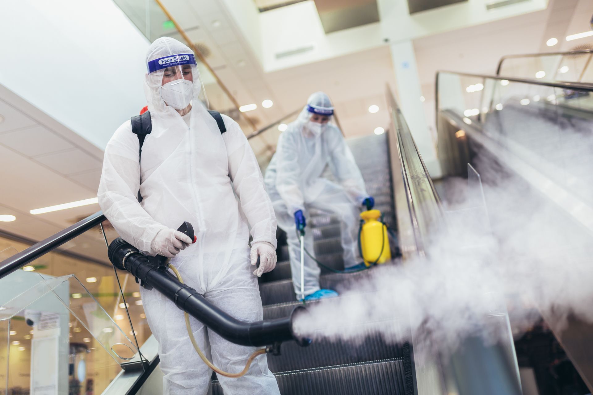 Two workers in protective suits fogging disinfectant in a mall corridor