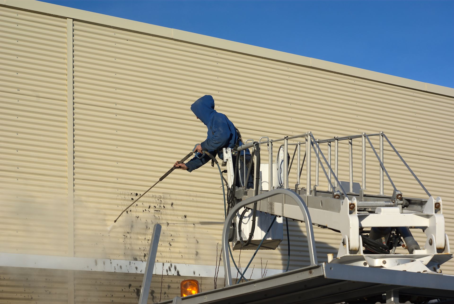 Worker in a blue hoodie pressure-washing a beige building wall from a raised platform.