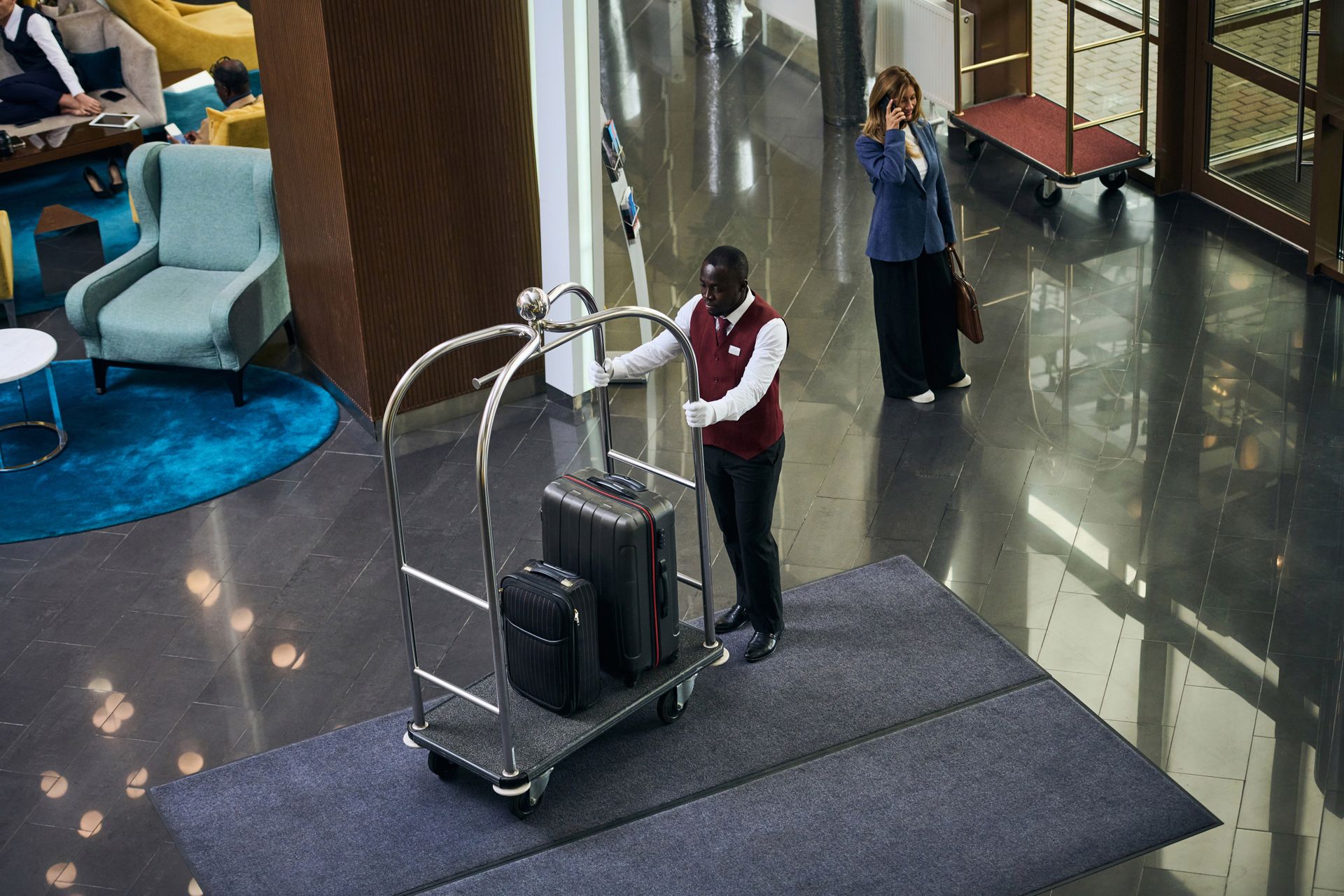 Hotel lobby with a bellhop pushing luggage cart, and a woman standing nearby on the polished floor.