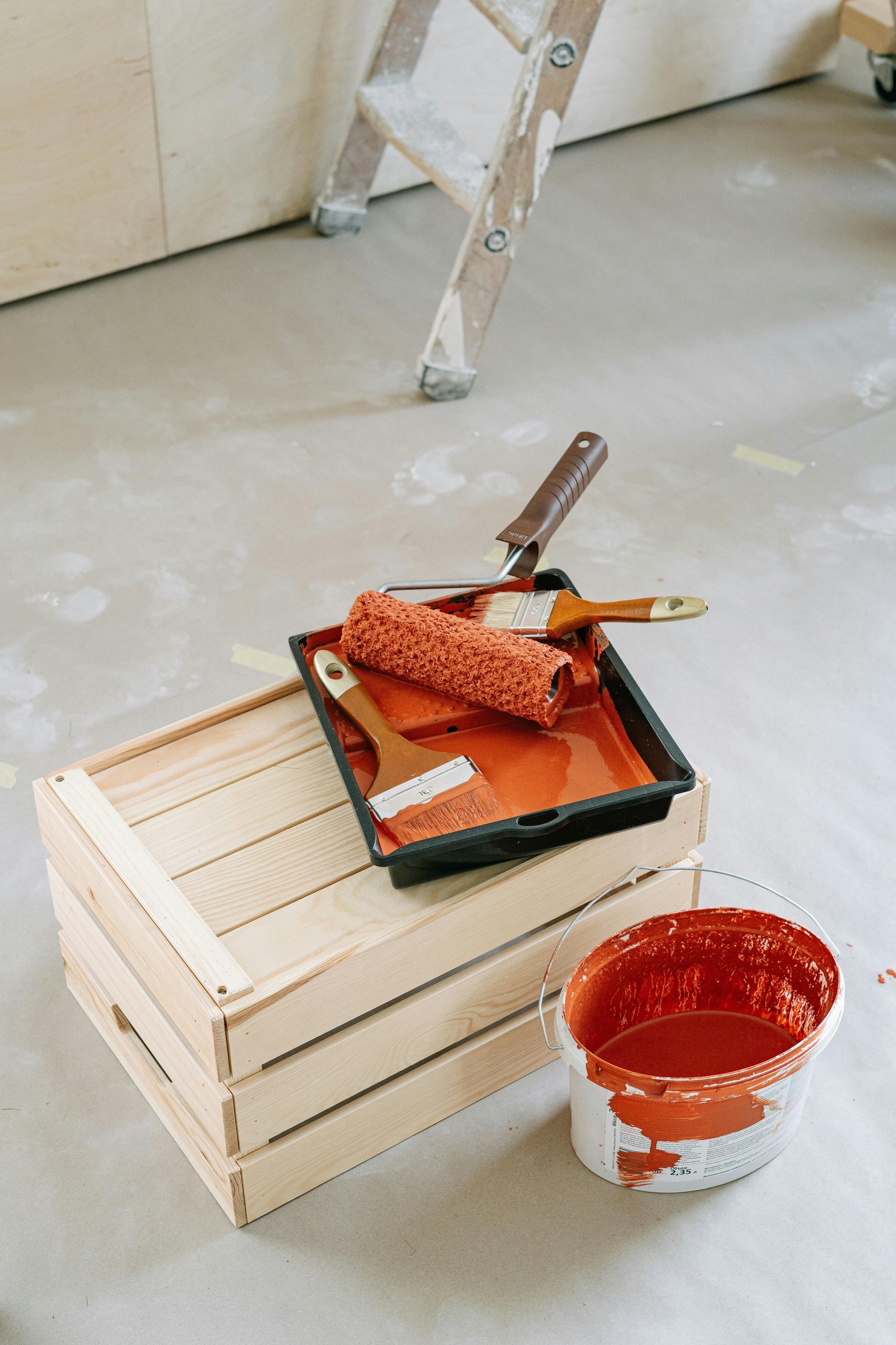 Paint supplies: open paint can and tray with roller and brushes on a wooden crate, with a ladder in the background.