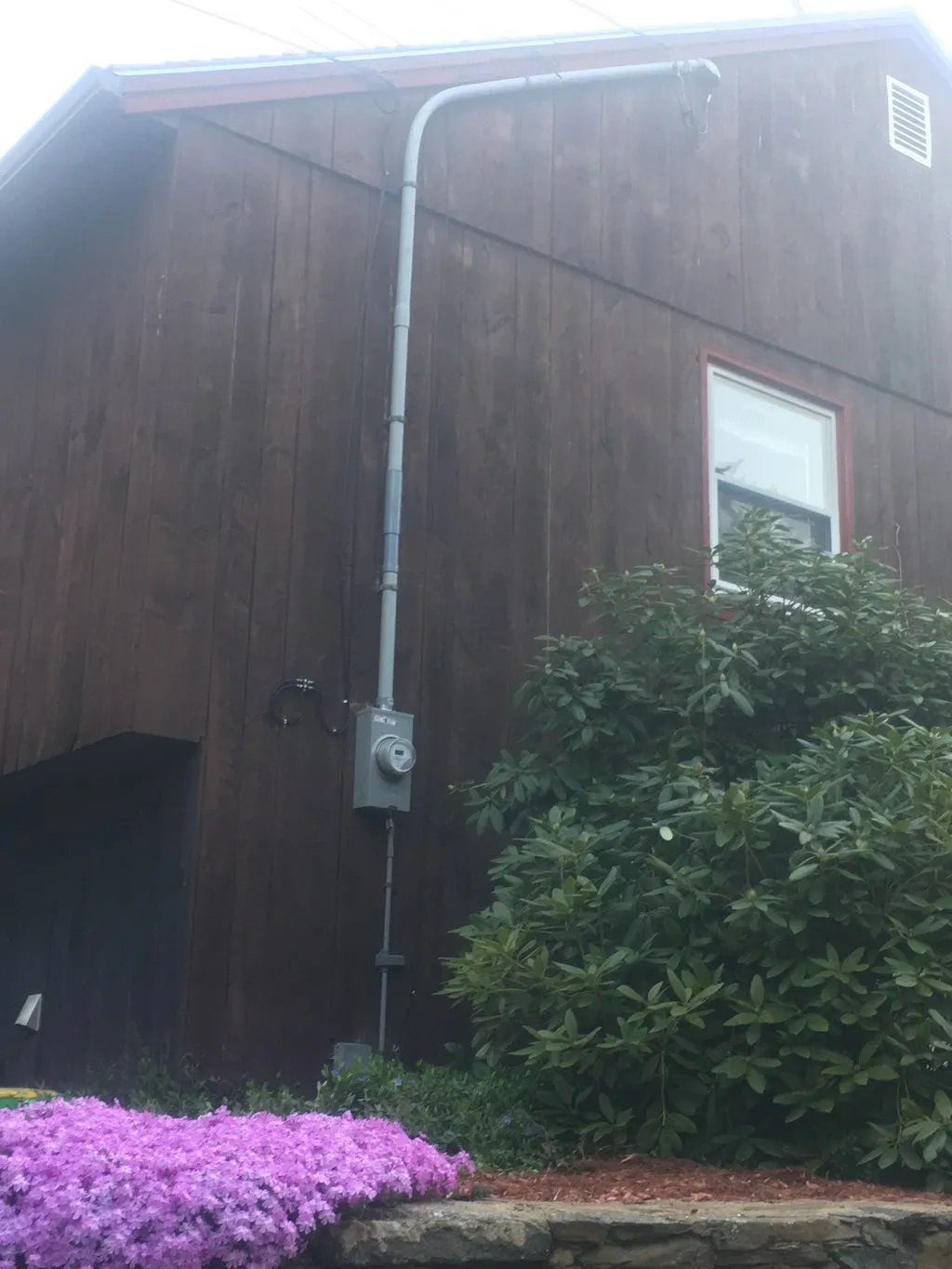 Electrical conduit and meter box on a brown wooden building exterior, near a window and greenery.