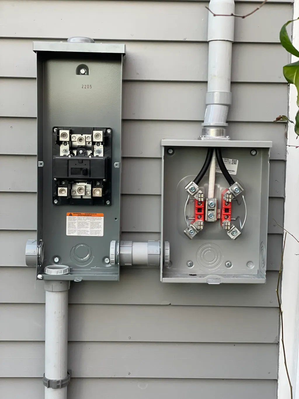 Metal electrical equipment on gray siding. Conduit pipes connect two boxes with exposed wiring.