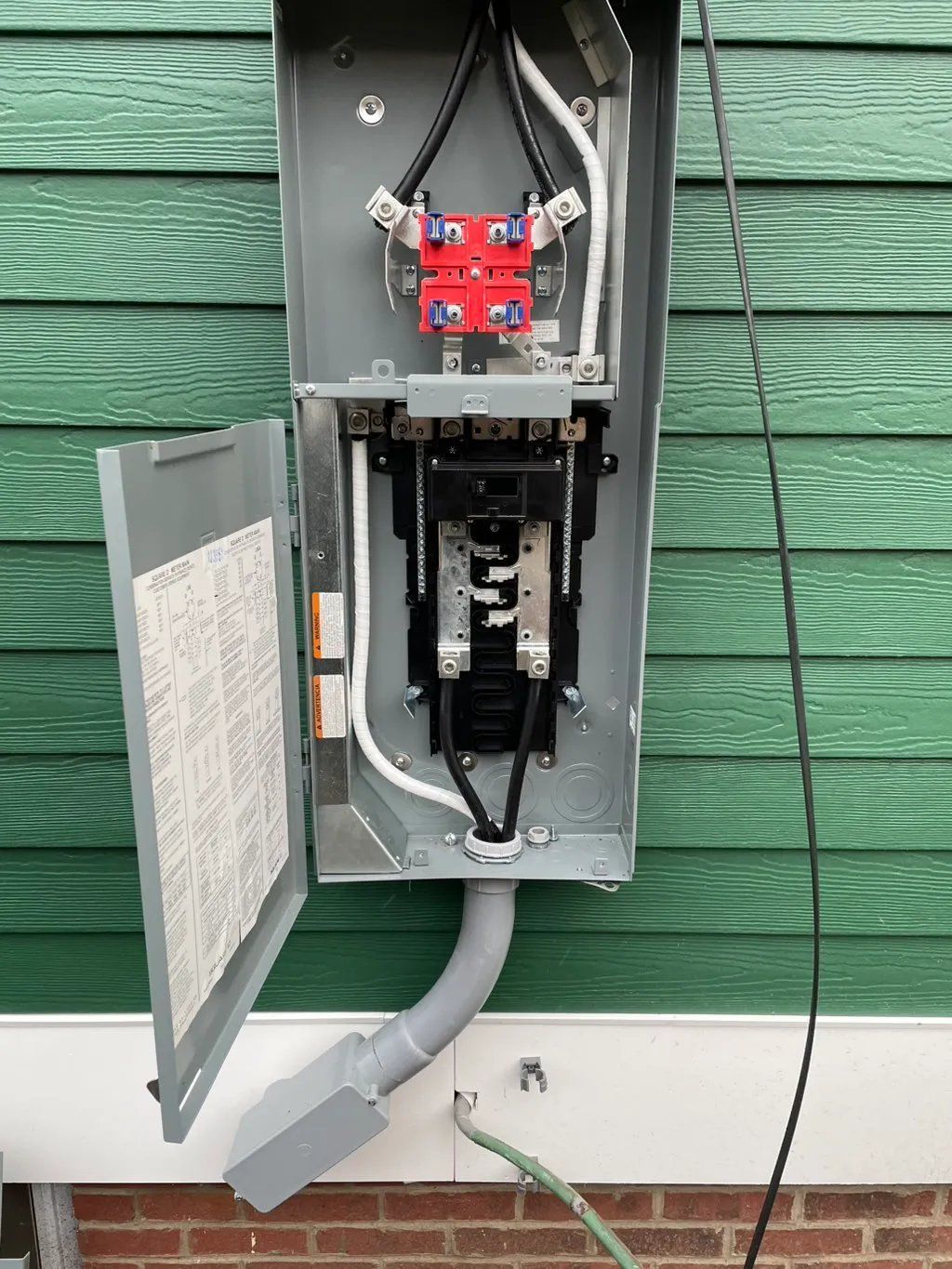Outdoor electrical panel with open door, green siding background. Gray metal box, wires, and components visible.