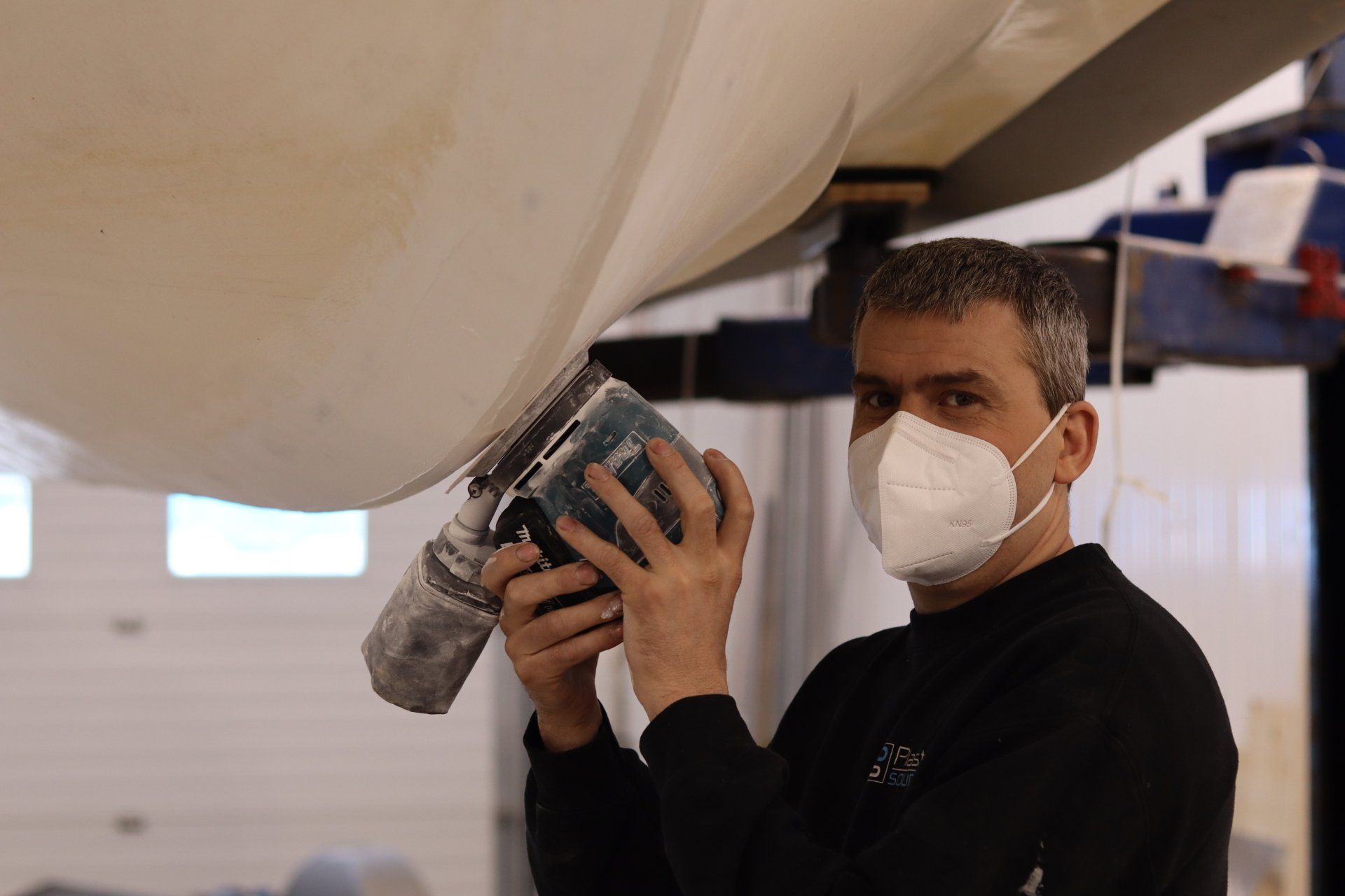 Un homme portant un masque utilise un outil électrique sous un bateau dans un atelier.