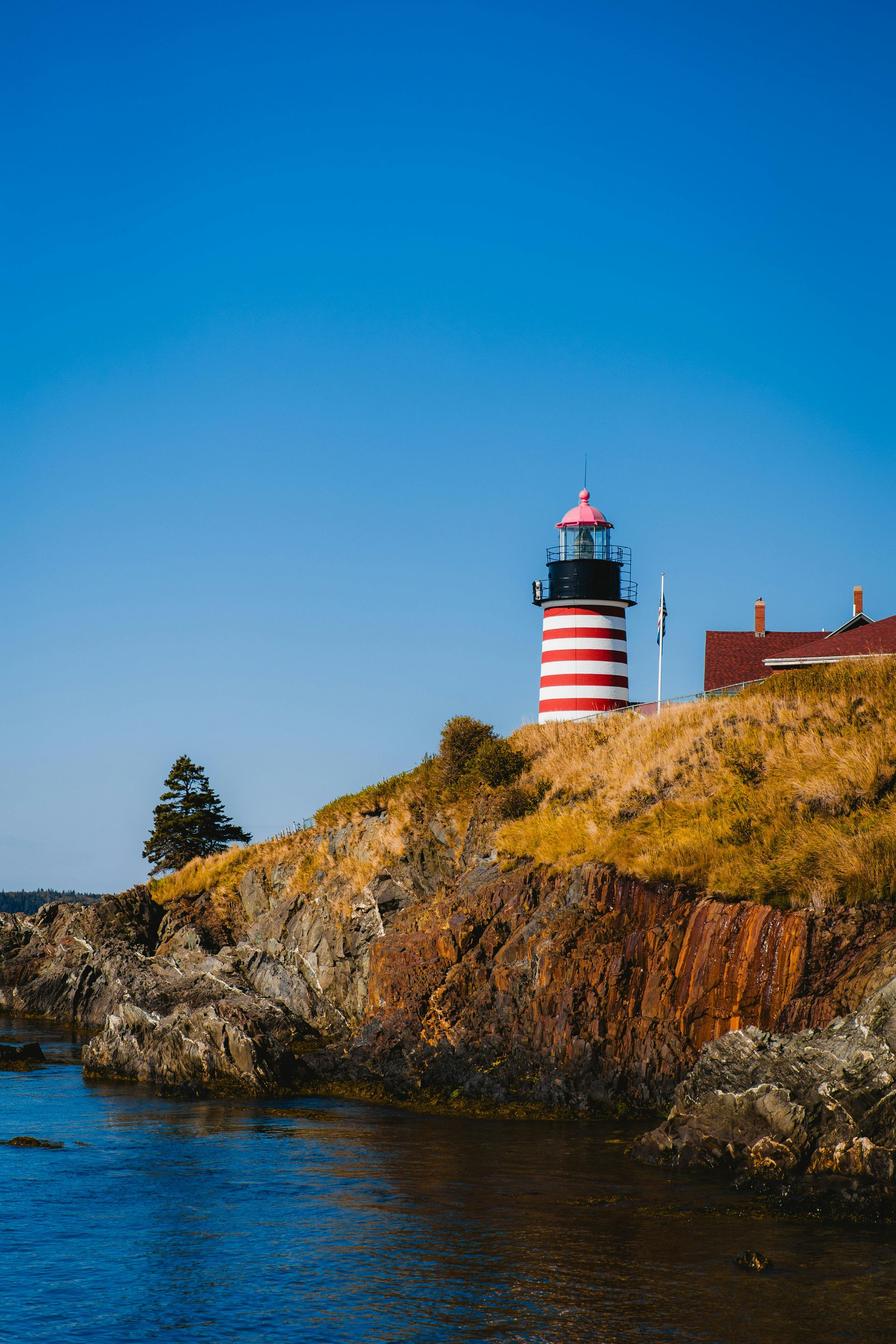 Phare à rayures rouges et blanches sur une falaise rocheuse, eau et ciel bleus.