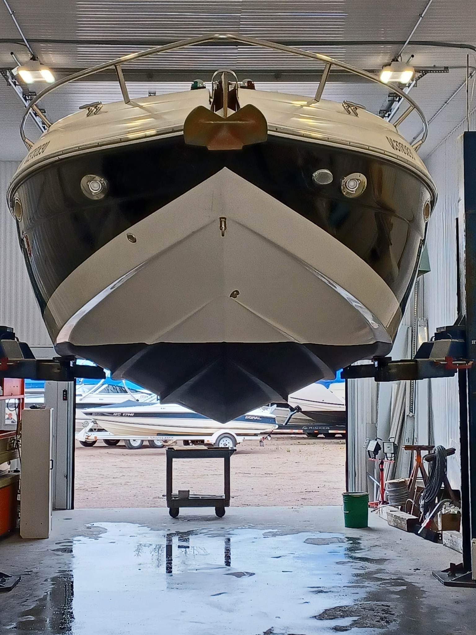 Coque de bateau soulevée sur des ponts élévateurs dans un atelier de réparation, laissant apparaître sa face inférieure. Coque sombre, métal gris.