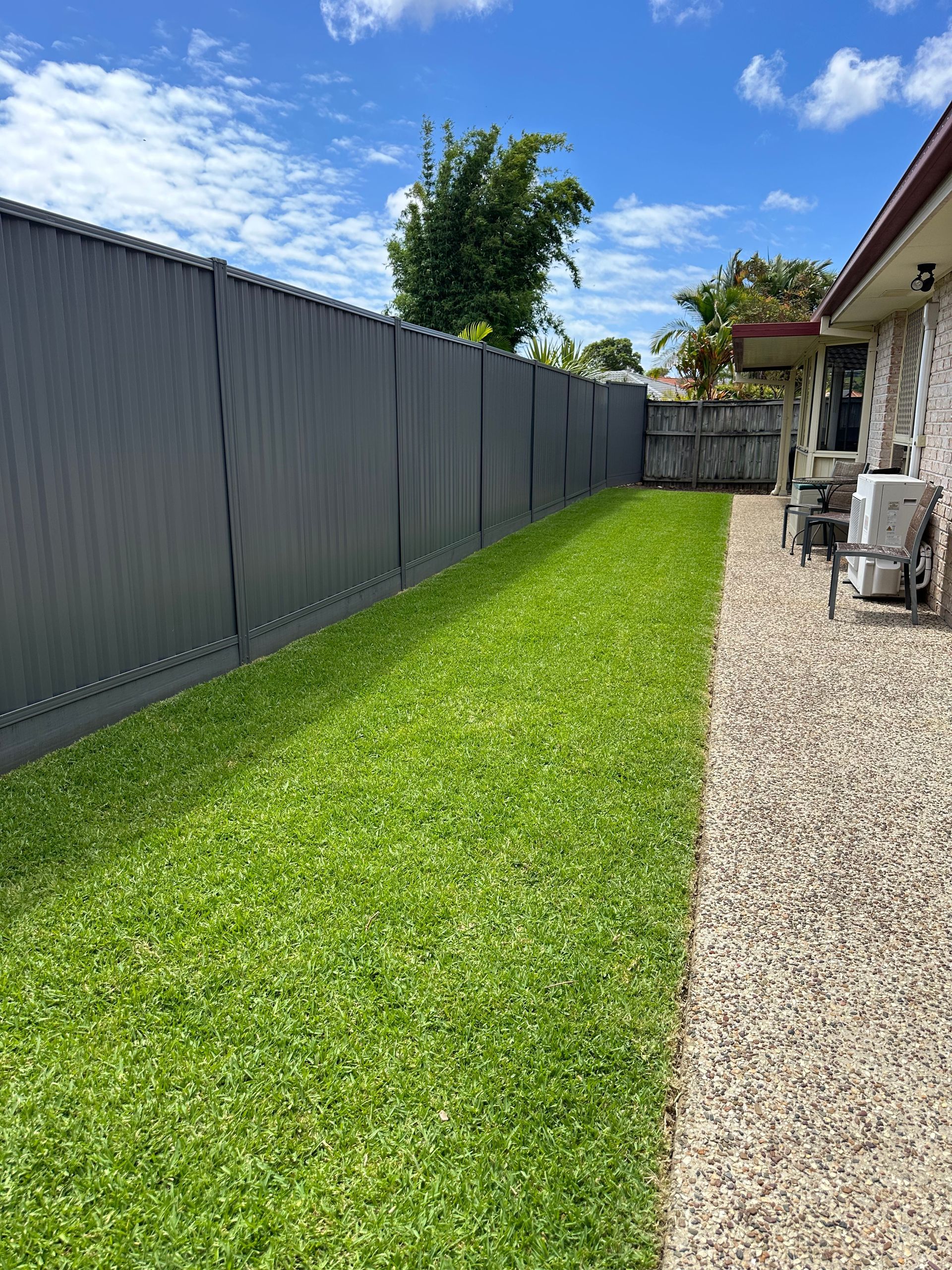 Lush green lawn along a colorbond fence line beside an exposed aggregate patio, leading to an outdoor seating area