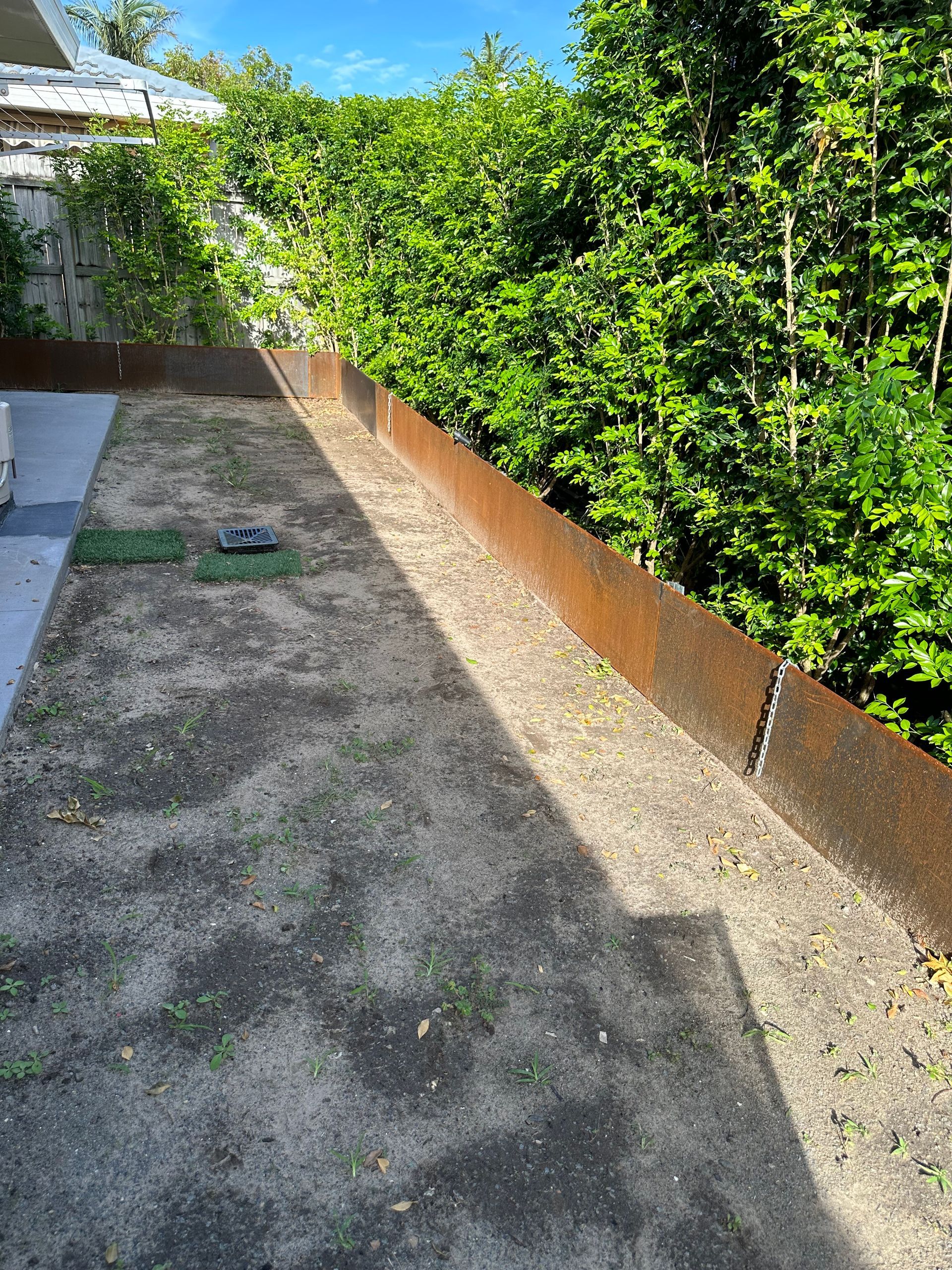 Straightcurve corten edging along a hedge boundary, with a stormwater drainage grate set into bare soil ready for turf
