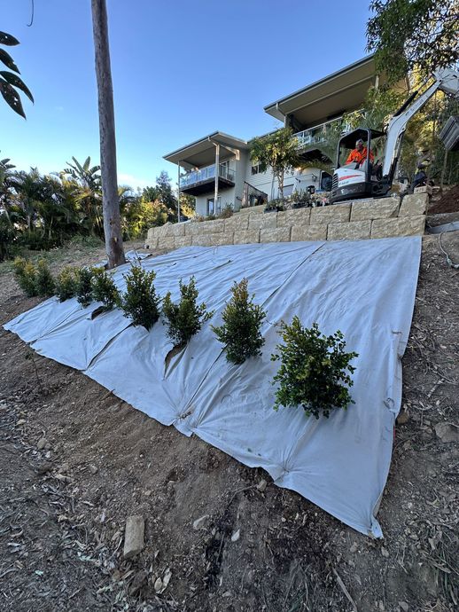 Completed sandstone retaining wall with excavator on site and native shrubs planted along a sloped garden bed