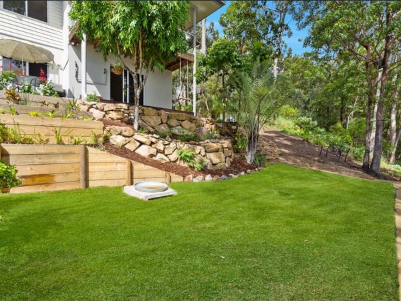 Tiered timber sleeper and natural rock retaining walls with garden planting and lush green turf, completed landscaping project on a sloped Gold Coast property