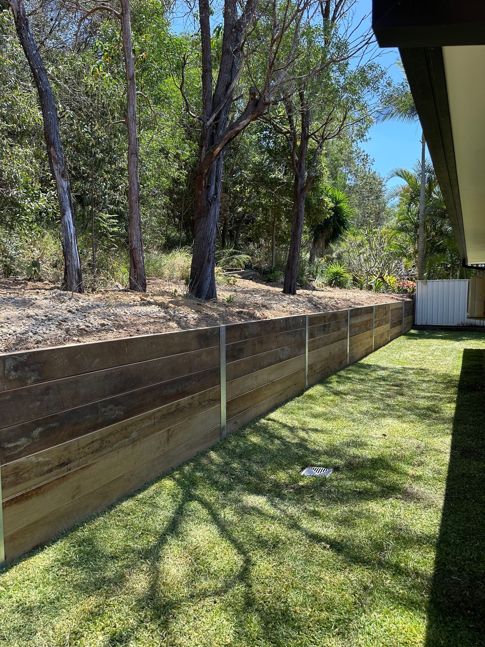 Timber sleeper retaining wall installation separating a landscaped lawn from natural bushland, with steel support posts and lush green grass in the foreground.
