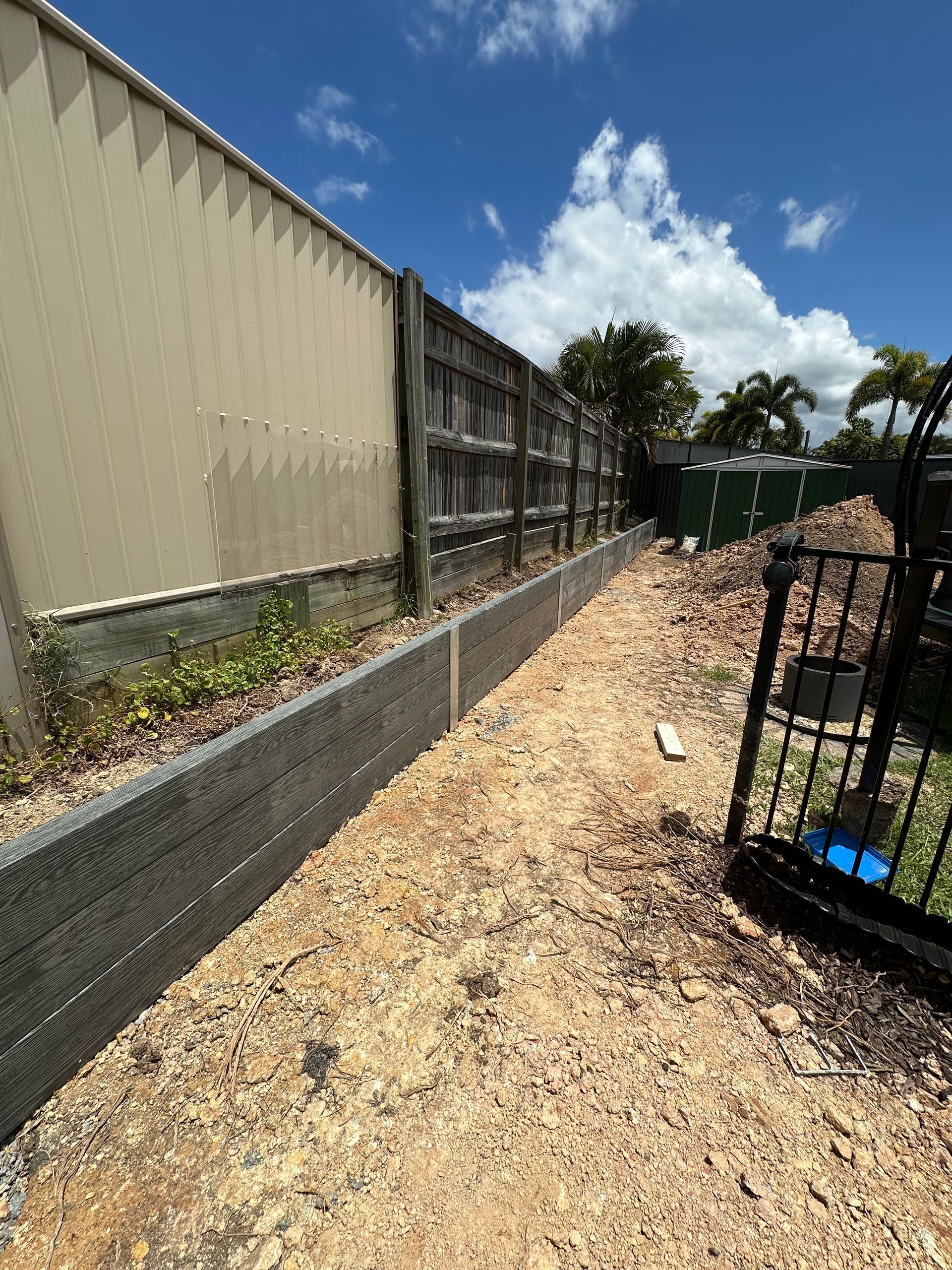 Tuffpoly composite sleeper retaining wall along a narrow side yard boundary next to a colorbond shed, mid-construction with excavated soil ready for backfill
