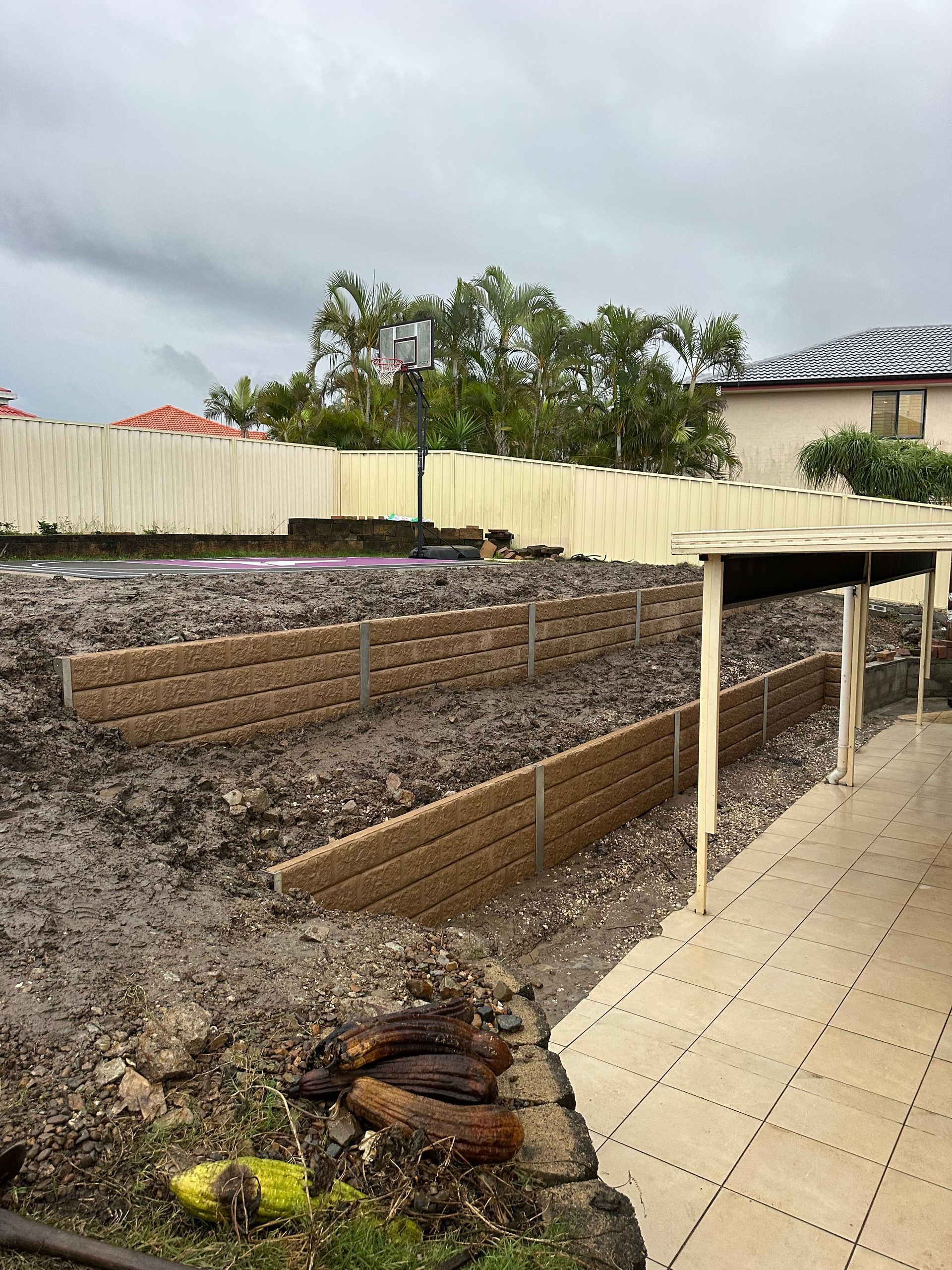 Tiered concrete sleeper retaining walls under construction in a Gold Coast backyard, levelling a sloped block near a home and basketball court