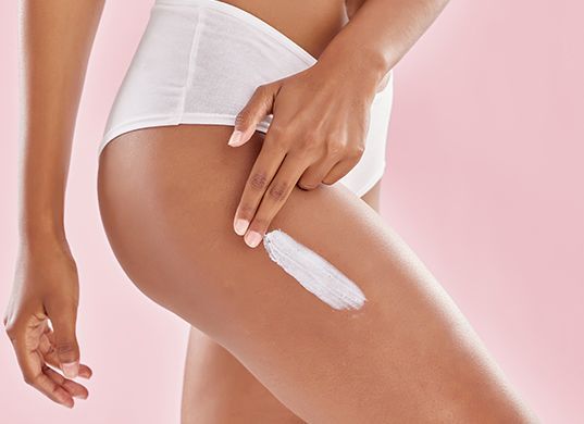 Person trägt weiße Lotion auf den Oberschenkel auf (vor rosa Hintergrund)