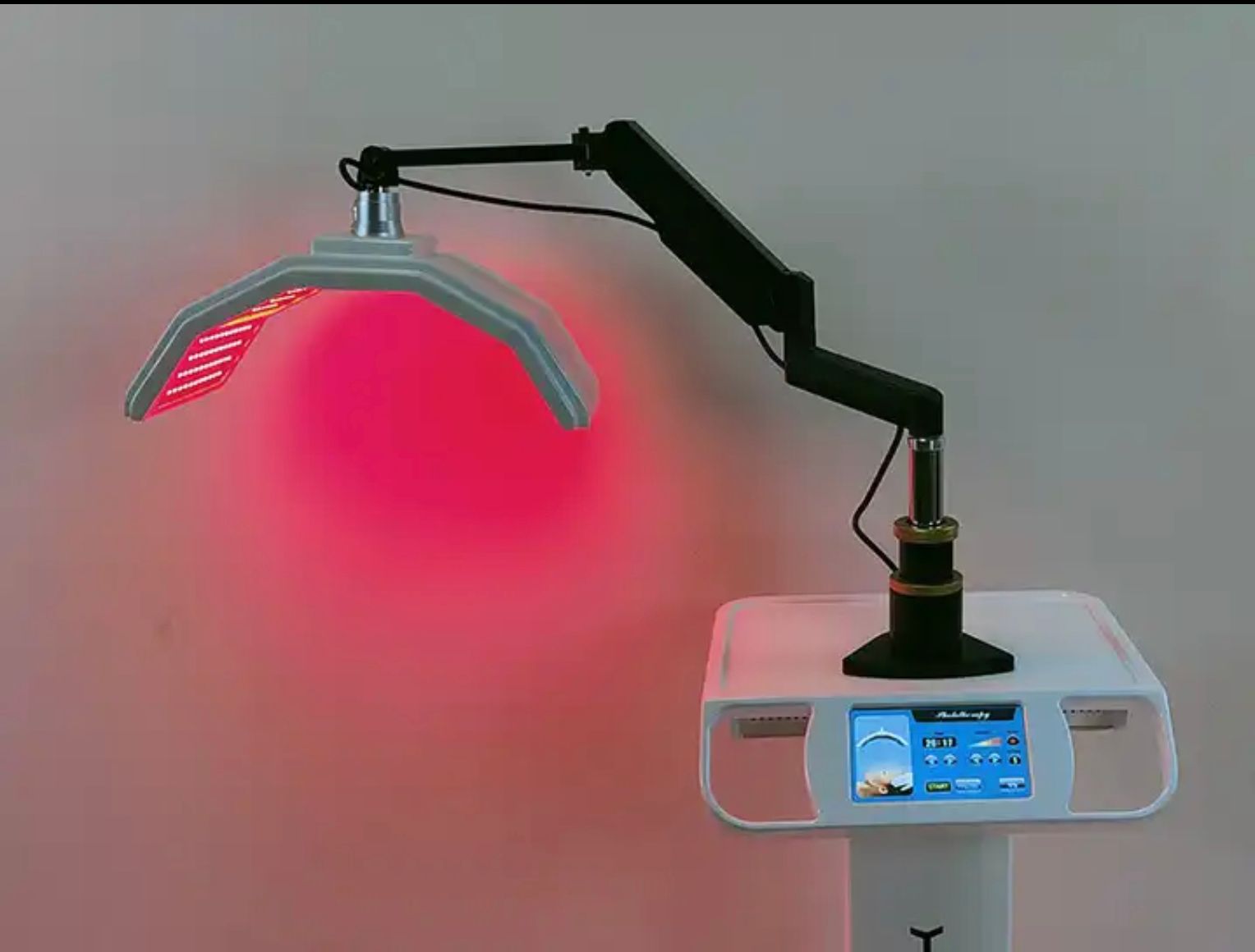 Eine Rotlichttherapielampe an einem verstellbaren Arm über einer Steuereinheit leuchtet rosarot vor einer grauen Wand.