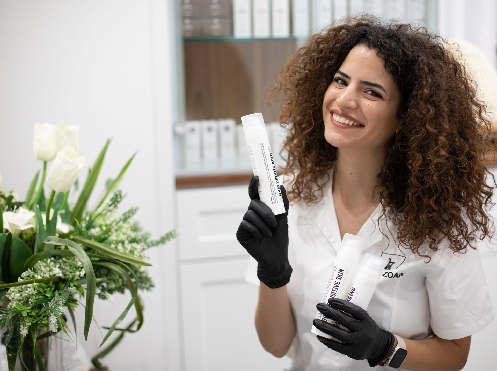 Eine lächelnde Floristin in weißer Uniform und schwarzen Handschuhen hält zwei Blumenstiele in einem Geschäft.