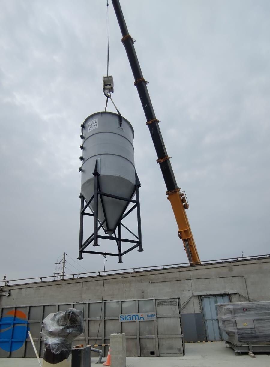 Una grúa eleva un gran tanque silo gris hasta la azotea de un edificio bajo un cielo nublado.