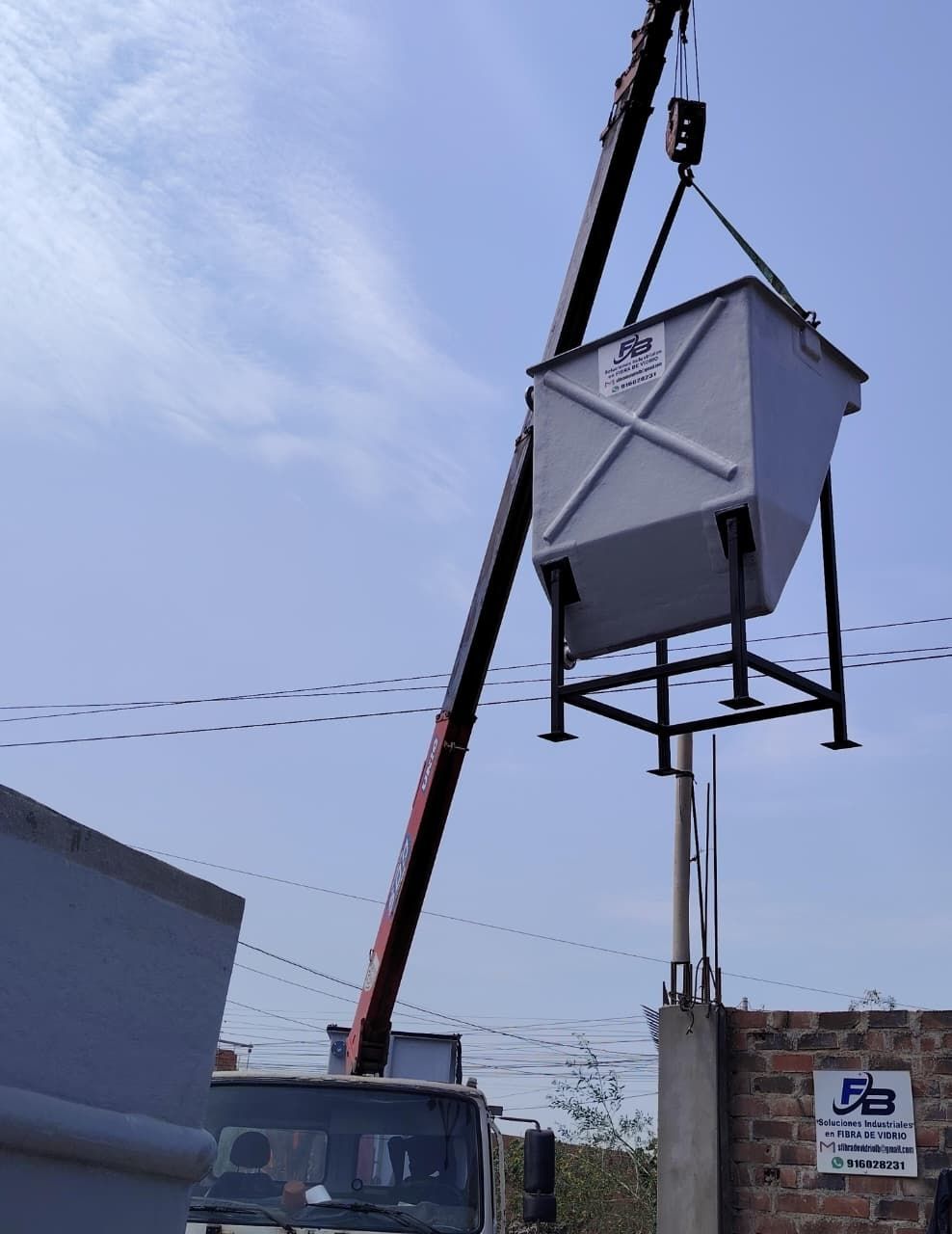 Grúa levantando una gran tolva metálica en una obra de construcción bajo un cielo azul.