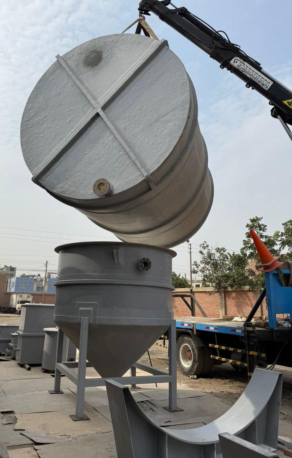Grúa de construcción levantando un gran tanque de metal gris en una obra.