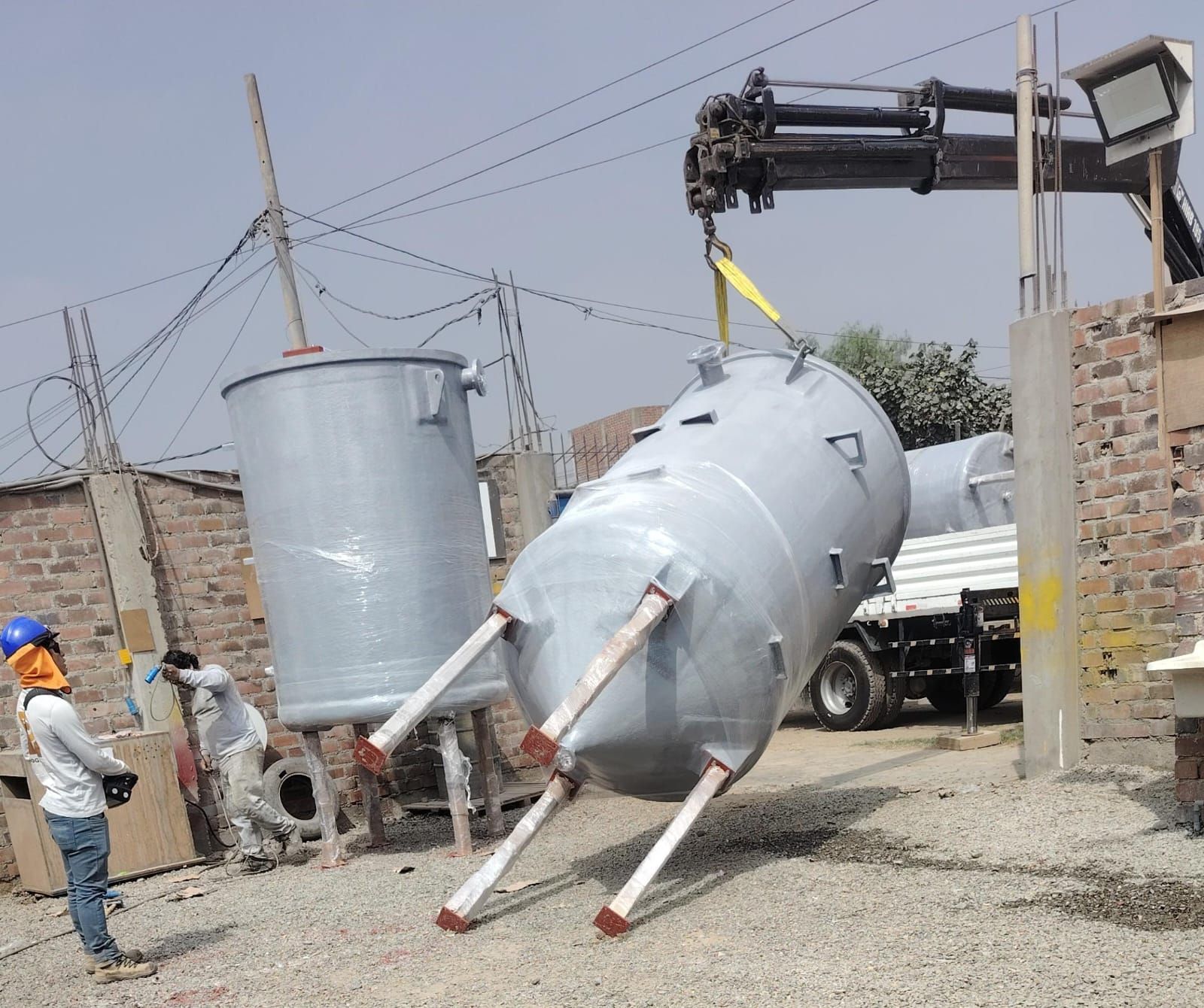 En un patio industrial al aire libre, unos trabajadores mueven un gran tanque cilíndrico gris con una grúa.